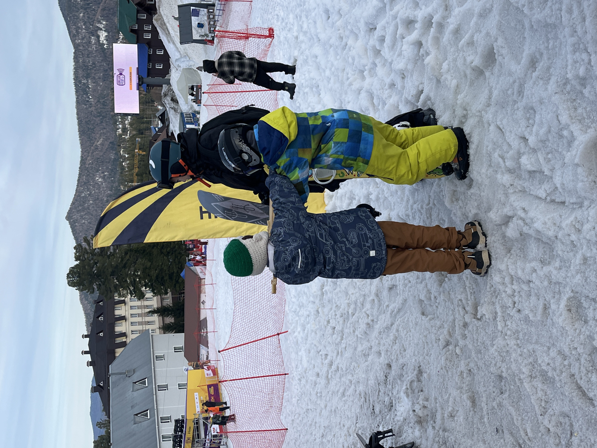 The image depicts a snowy scene, likely at a ski resort, with three people in the foreground. Two are snowboarders wearing helmets and winter attire, and they appear to be adjusting their gear or interacting with each other. The third person is a child wearing a patterned jacket, brown pants, and a green and white knit hat, possibly observing the snowboarders.
Behind them, a red safety net separates the foreground from a more populated area. There is a large screen displaying some event branding. Buildings are visible in the background, along with a forested hillside and a mountain range in the distance. The location appears to be a ski resort in a mountainous area.
