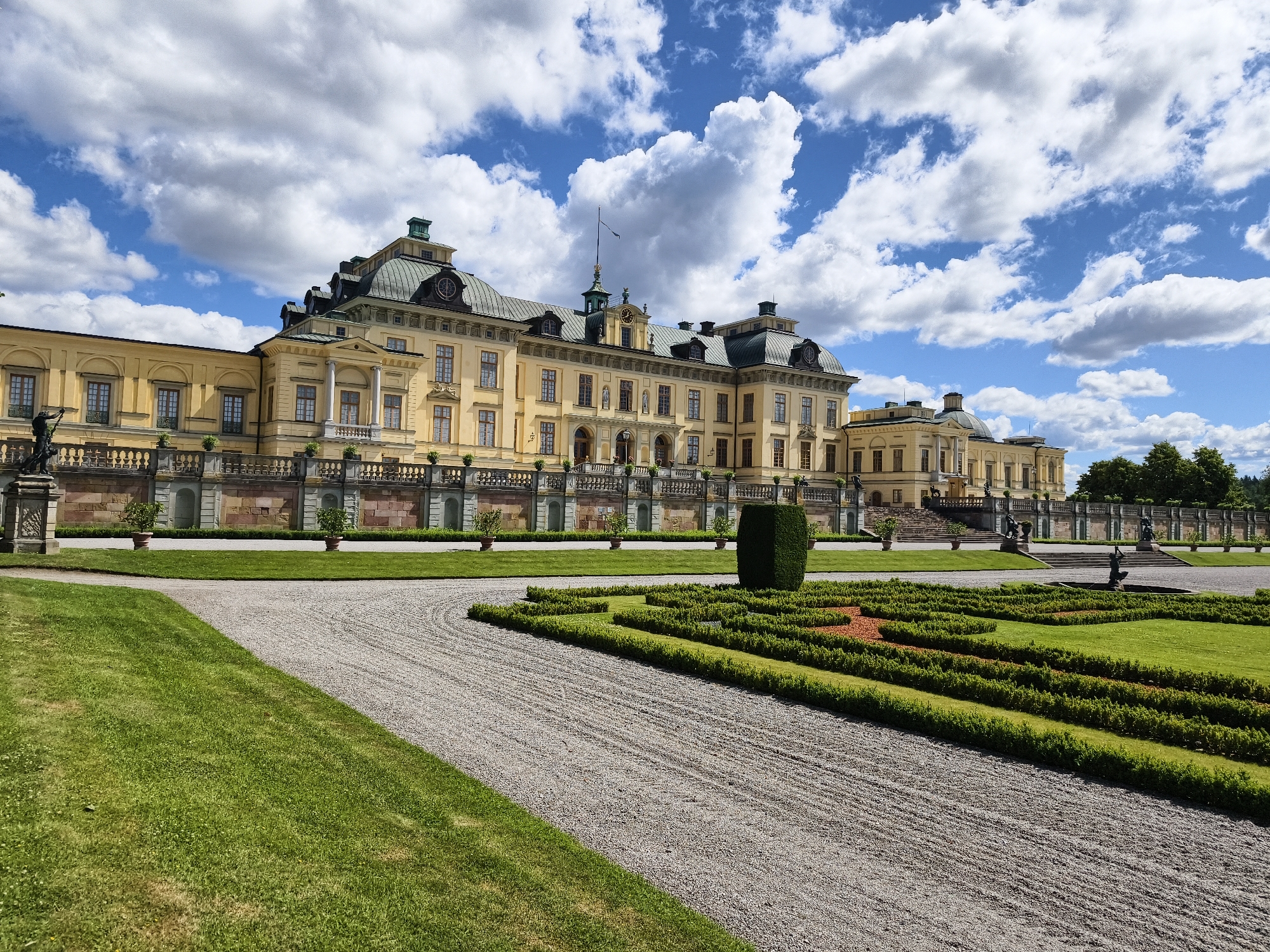 This is an outdoor, eye-level, wide-angle photograph of the exterior of the Drottningholm Palace in Ekerö kommun, Sweden. The palace, painted a pale yellow, is a large, baroque-style building with multiple wings, domes, and classical architectural details. It is set against a bright blue sky filled with large, fluffy white clouds. In the foreground, there is a meticulously maintained garden with manicured green hedges forming geometric patterns, interspersed with patches of reddish-brown soil. A gravel pathway curves through the garden. To the left of the gravel path is a vibrant green lawn. A tall, dark green, rectangular hedge stands prominently in the middle-right of the garden. Behind the garden, a stone wall with decorative balustrades stretches across the width of the palace. Statues and decorative urns are visible along the wall and on the palace's terraced entrance. The overall scene depicts a grand architectural structure surrounded by formal gardens under a sunny, cloudy sky.