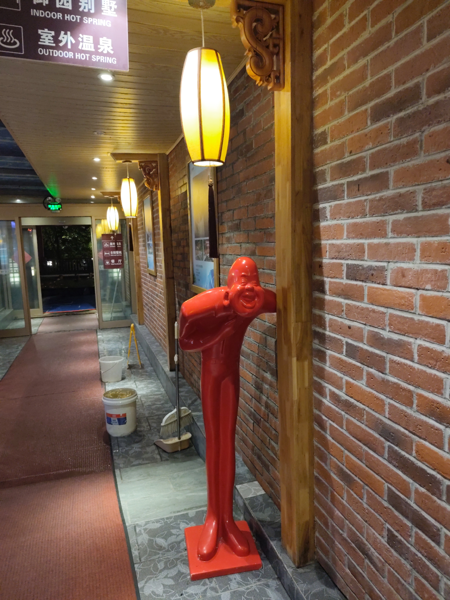 The image captures a scene outside a building with a prominent brick wall. In the foreground, a bright red, stylized human figure, possibly a statue or a prop, stands leaning against a wooden beam. The figure has a distorted, smiling face and outstretched arms. To the left of the figure, a red carpet runs along a pathway leading towards an open doorway with glass doors. Inside the building, through the glass doors, a dimly lit hallway with a blue floor can be seen, along with several signs.
The brick wall is made of reddish-brown bricks with visible mortar. There are two large, lantern-like light fixtures hanging from the ceiling, illuminating the area with a warm yellow glow. Next to the red figure, on the tiled ground, is a broom and dustpan, and two white buckets, one of which appears to contain some yellow substance.
Above the doorway, a sign with Chinese characters and English text reads "INDOOR HOT SPRING" and "OUTDOOR HOT SPRING," suggesting the location is a hot spring facility in 林甸镇, China. The overall environment appears to be an entrance or pathway area of a commercial establishment, possibly illuminated for evening or night.