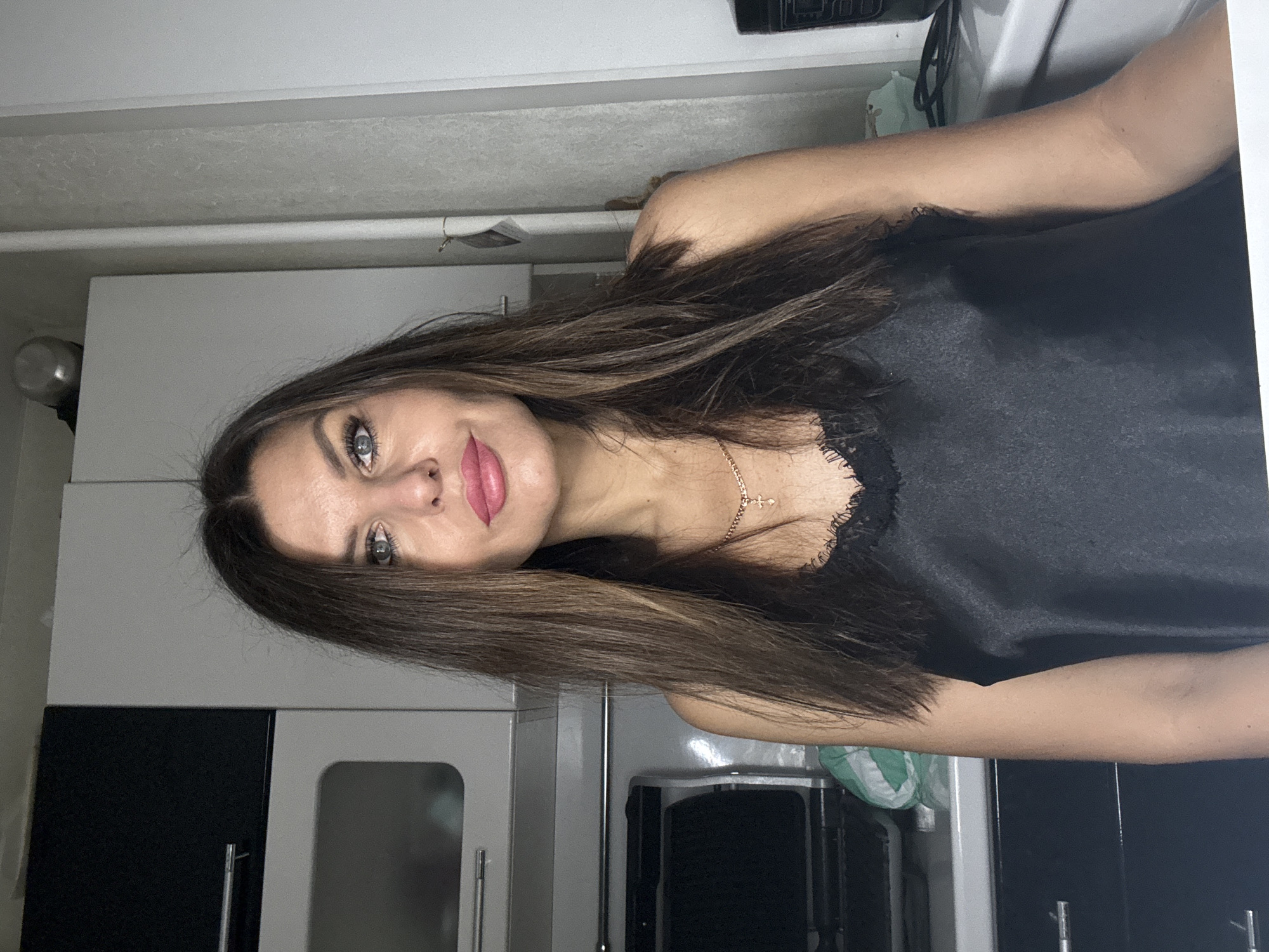 A woman with long brown hair and blue eyes is indoors. She is wearing a black top with lace trim and a gold necklace with a cross. The background appears to be a kitchen with white and black cabinets. The photograph was taken at a close range.