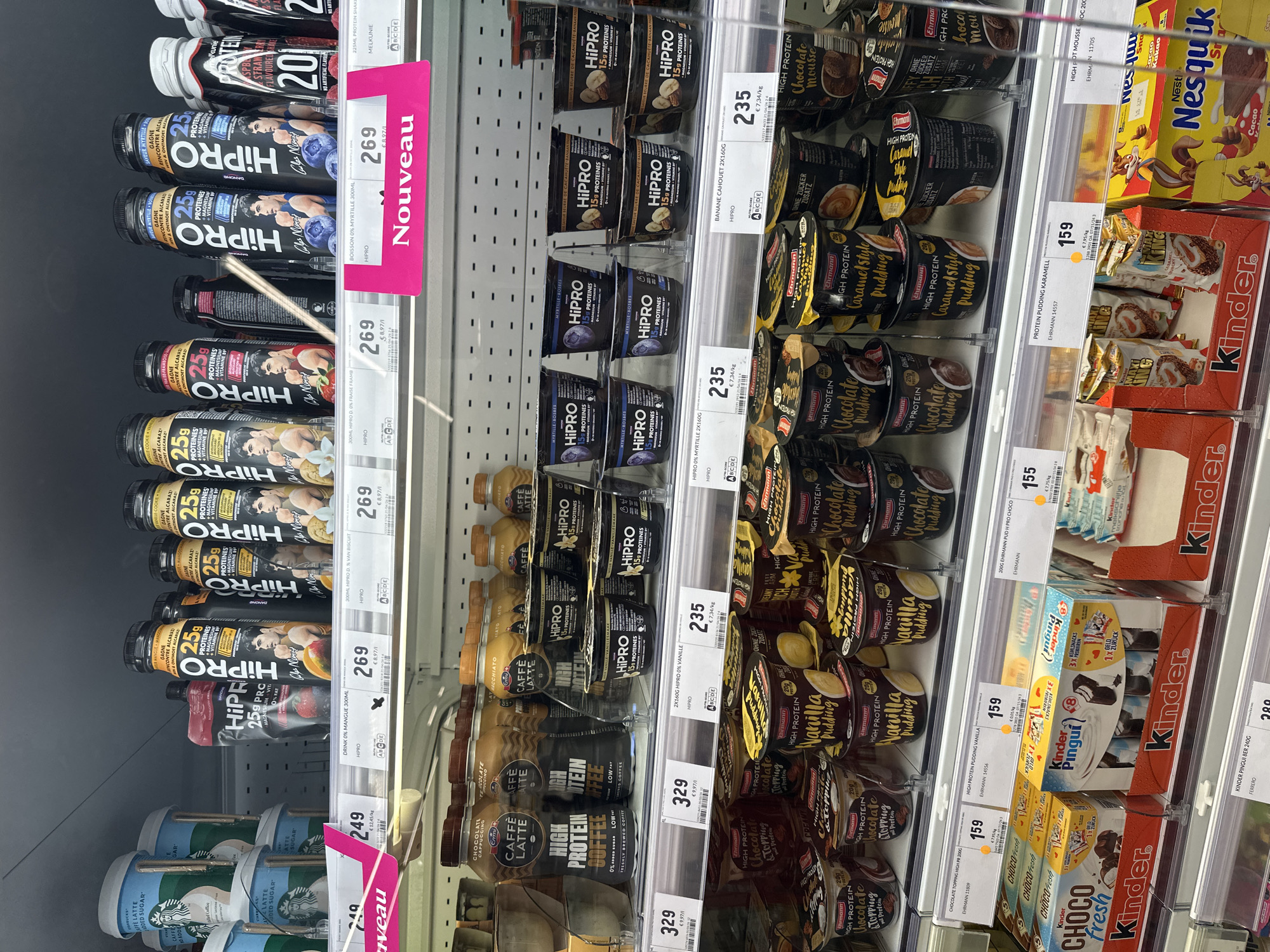 The image shows a refrigerated display case in a grocery store, likely a supermarket, in Echternach, Luxembourg. The shelves are filled with various food and drink items, primarily focusing on high-protein products and desserts.
On the top shelf, there are several bottles of "HiPRO" protein drinks in different flavors, including "0% mangue" (0% mango) and "0% d. van biscuit" (0% vanilla biscuit). These bottles are black with prominent "HiPRO" branding and images of men. There are also some "Starbucks Cafe Latte" cups visible on the far left.
The middle shelves are stocked with "HiPRO" yogurt cups, featuring flavors like "Myrtille-Bosses" (Blueberry) and "Beurre de cacahuète-banane" (Peanut butter-banana). Below these, there are bottles of "CAFFE LATTE HIGH PROTEIN COFFEE" and more "HiPRO" protein drinks.
The bottom shelves display a variety of desserts, including "Ehrlmann High Protein Chocolate Topping," "High Protein Vanilla Pudding," and "High Protein Caramel Style Pudding." Further to the right, there are boxes of "Kinder Choco Fresh," "Kinder Pingui," and "Nestle Nesquik Cacao."
Price tags are visible for many of the products, indicating prices in Euros. Some of the labels also show weight and nutritional information. A prominent pink "Nouveau" (New) sign is attached to one of the shelves, highlighting new products. The overall environment is a well-lit, organized retail setting, typical of a supermarket's refrigerated section. No people are visible in the image.