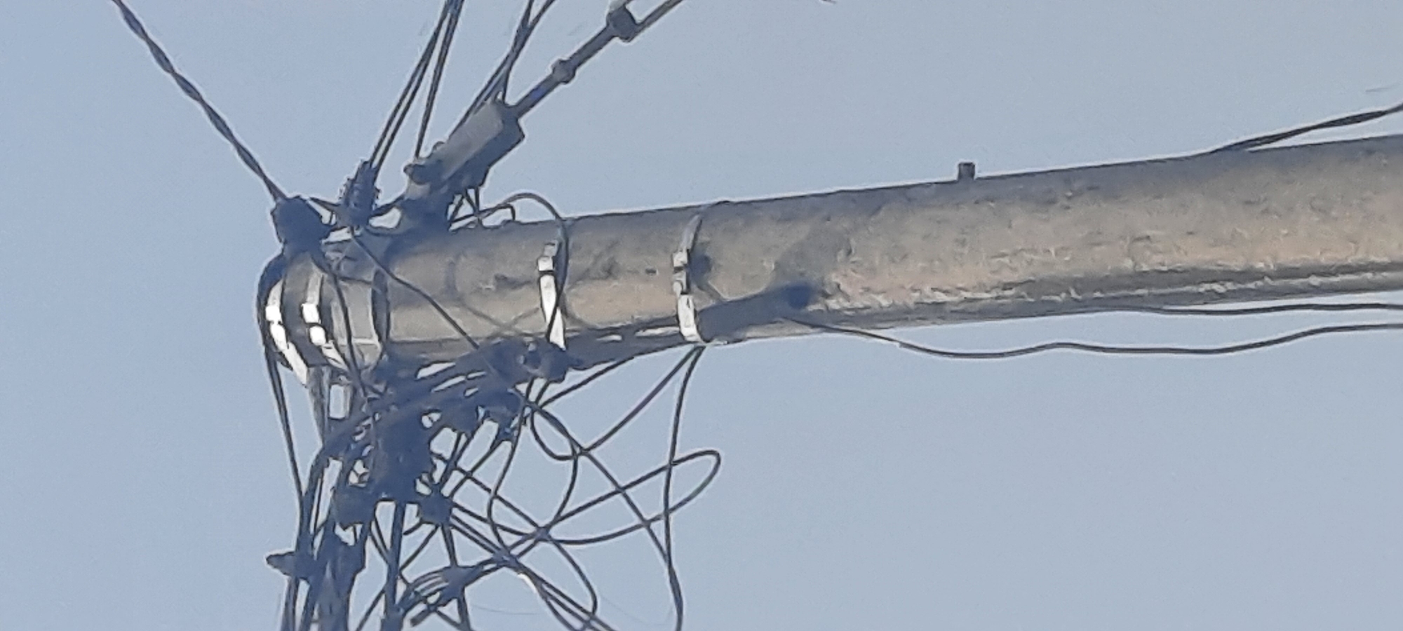 A concrete utility pole stands against a light blue sky, laden with a chaotic tangle of black wires. The pole itself is weathered, with visible texture and some grime. Two metal bands are cinched around the pole at different heights, likely to secure various components. The wires are numerous and complex, some appearing to be twisted together, while others hang loosely. They are attached to various points on the pole and extend outwards and upwards, disappearing out of frame. The scene is characteristic of urban or suburban utility infrastructure, suggesting a location where electrical and telecommunication lines are managed overhead. It is a photograph taken from below, looking up at the pole and its extensive wiring. The location is Ploso, Indonesia.
