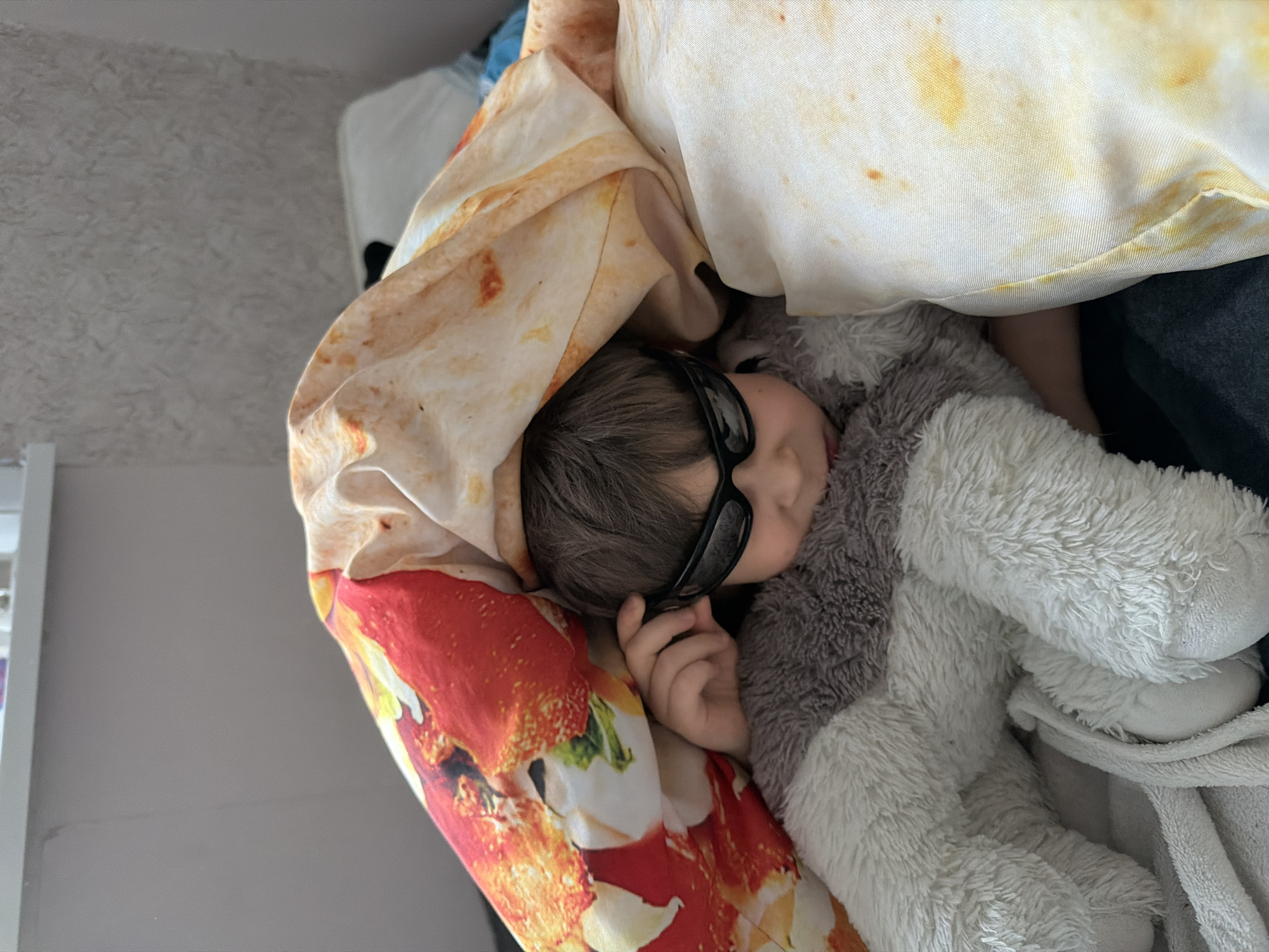 A young child wearing black sunglasses is lying down, wrapped in a pizza-printed blanket, and cuddling a gray and white stuffed animal. The setting appears to be indoors, possibly in a bedroom or living room. The location is specified as Perm, Russia.