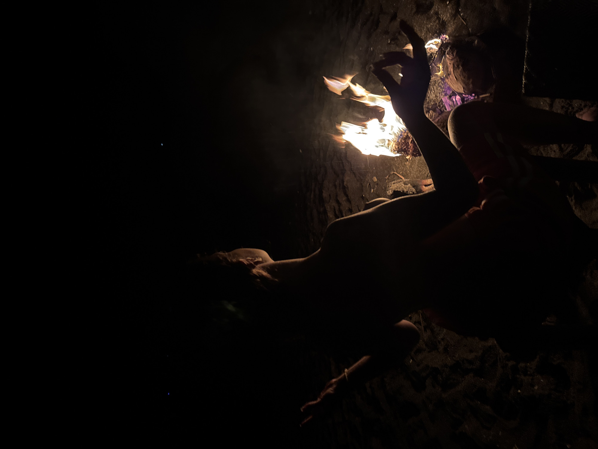 Two people are sitting near a fire on what appears to be a beach in the dark. One person has their arm raised near the fire. The environment is dark.