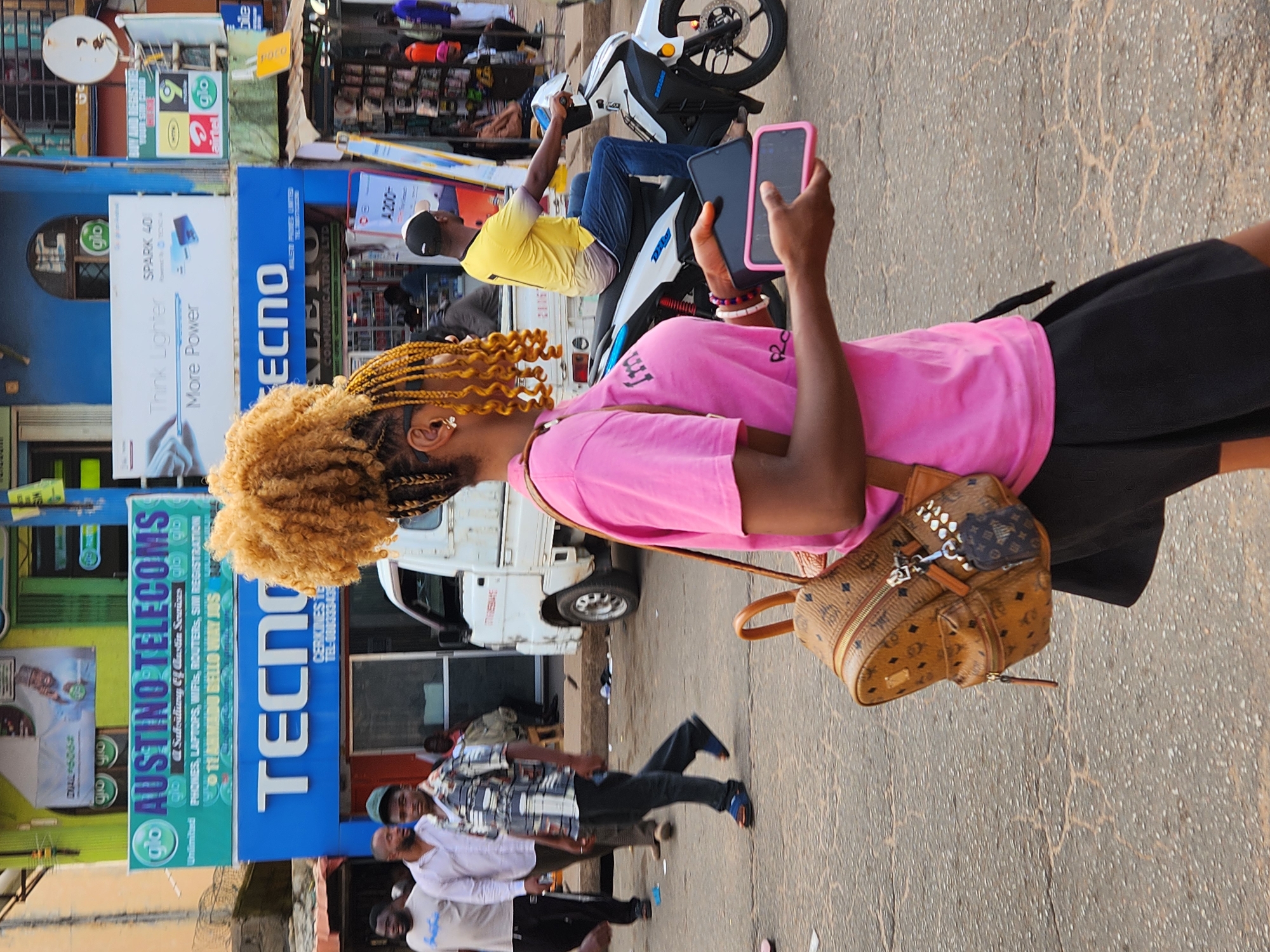 The scene is a bustling urban street in Jos, Nigeria. A woman with blond braids and a pink shirt is prominently featured, holding a pink-cased smartphone. A man on a white motorcycle behind her also holds a phone. Several people are walking in the background. Blue and green signs for "Austino Telecoms" and "TECNO" are displayed on the buildings, promoting phones, laptops, and other services. A white truck is also visible. The street surface is paved.