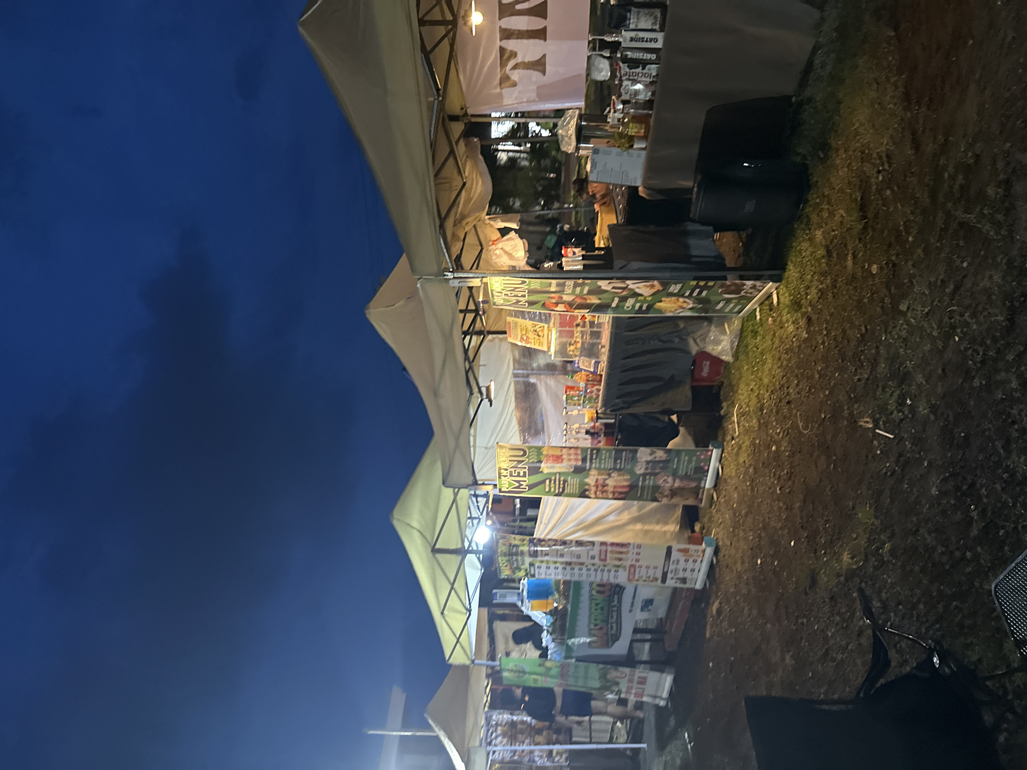This image captures an outdoor night market or food festival in Davao City, Philippines. The scene is dominated by a row of temporary vendor stalls, each covered with a light-colored canopy, likely white or beige. Artificial lighting emanates from within and around the stalls, illuminating the immediate area against the dark blue night sky, which appears clear.
The ground beneath and in front of the stalls is unpaved and covered with sparse, dry grass. Various banners and signs are displayed on the stalls, showcasing menus and food items. Notable text includes "MENU" on green banners, with one clearly indicating "BURGER" (likely "BURGER MENU"). Another prominent sign reads "MASTERBOOST." Boxes labeled "OATSIDE" are stacked on one of the tables, and partial text like "MIL" and "Jacgato" (or similar) can also be seen on banners.
A large, dark object, possibly a speaker (with "JBL" brand visible), rests on the grassy ground near the right side of the image, suggesting music or announcements. A black mesh chair is partially visible in the bottom right corner. Although the market appears to be operational, few people are distinctly visible; most are either obscured by the stalls or present as indistinct figures in the background. The overall impression is that of a bustling but casually set-up evening event focused on food and beverages.