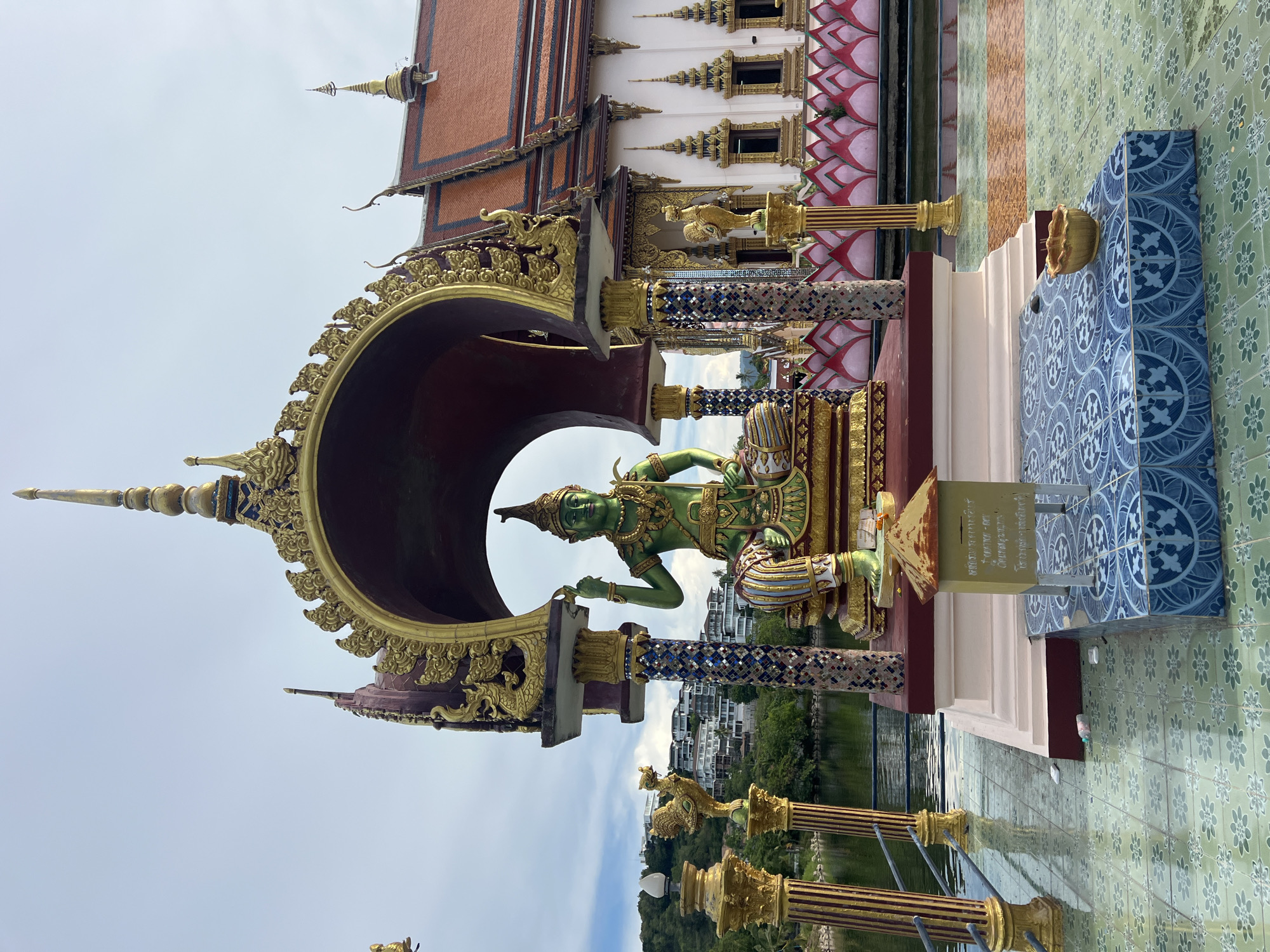 The image captures a serene outdoor scene at the Baan Plai Laem Temple in Koh Samui, Thailand. Dominating the foreground is a vibrantly painted green statue of a deity, seated on a gilded platform. The deity is adorned in traditional attire, with intricate golden jewelry and a distinctive pointed headdress. The statue is housed within an ornate, arched structure, heavily decorated with golden, swirling motifs and supported by colorful, mosaic-tiled pillars.
To the right of the statue, the temple itself presents a striking architectural facade with a red tiled roof, adorned with decorative spires and intricate golden trim. A series of lotus-like pink and white decorative elements line the base of the temple wall, situated along the edge of a calm body of water.
In front of the deity's platform, a small, weathered donation box with a pointed roof sits on a blue, patterned tiled surface. A golden incense burner rests on the same tiled area. Scattered on the tiled floor in the midground are a few pieces of litter, including a plastic bottle and some small white objects.
To the left of the statue, a golden, ornate bird statue, possibly a mythical creature like a garuda, stands on a golden pedestal. Beyond this, a calm body of water reflects the overcast sky. In the background, a lush green hillside is dotted with modern residential buildings, suggesting the temple's location in a developed area. The overall atmosphere is one of religious devotion combined with natural beauty.