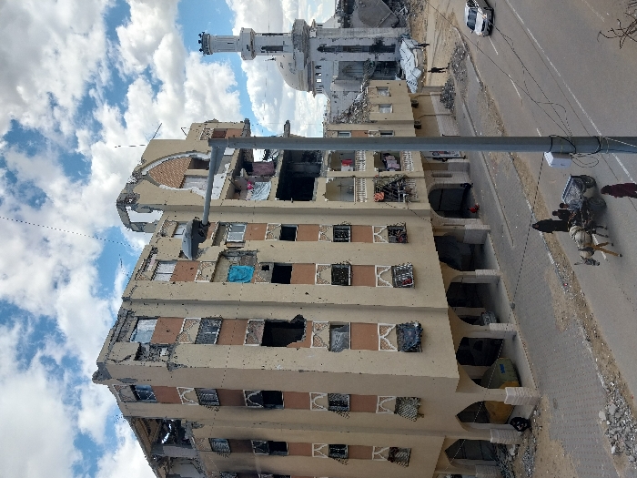 This image depicts a scene of extensive urban destruction in Khan Yunis, Palestinian Territory, under a partly cloudy sky.
The central focus is a multi-story residential or commercial building, approximately six to seven floors high, exhibiting severe structural damage. Its light-colored facade is heavily scarred with numerous impact marks, holes, and shattered windows. Large sections of the roof and upper floors are destroyed or collapsed, exposing interior rooms. Some windows are entirely blown out, while others show tattered coverings or debris. Despite the damage, laundry or clothing items are visible hanging from several balconies and windows, suggesting recent or continued presence. The ground floor features a series of arched openings, some appearing to be entrances.
To the right and further back, a mosque with a distinct minaret and dome is visible, also heavily damaged and partially collapsed, surrounded by rubble. Beyond the mosque, more extensively damaged structures are discernible.
In the foreground, a paved street runs left to right. Debris and rubble are scattered along the left sidewalk and road edge. A grey street light pole stands prominently in the mid-right. On the street, a donkey-drawn cart, carrying at least two individuals, is moving towards the right. Nearby, a person in dark clothing walks on the sidewalk, and another in reddish attire is closer to the cart. A silver sedan is visible driving away on the right lane of the street. Beneath the arches of the main building, two individuals are seated or resting on the ground.