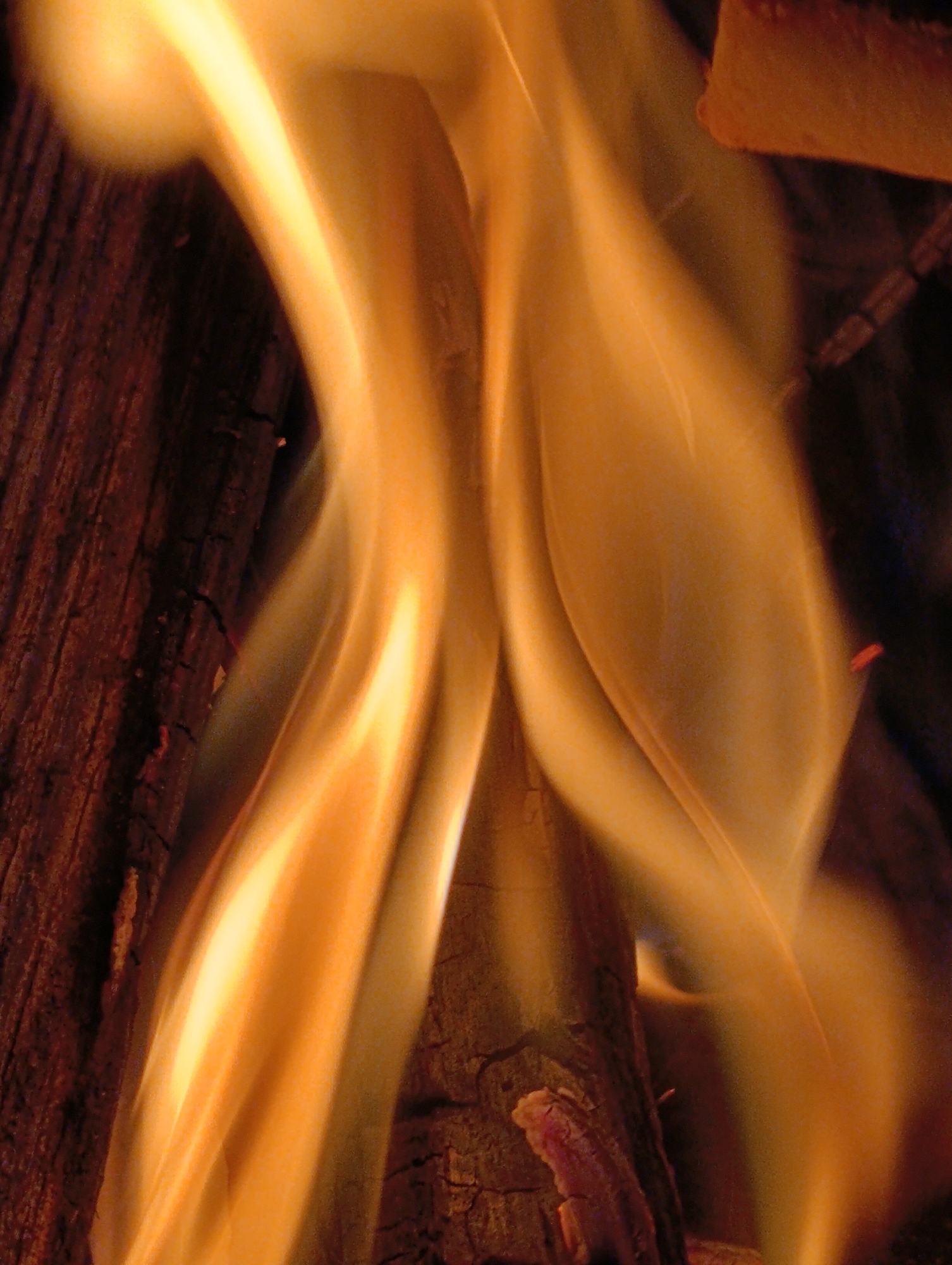 This is a close-up, eye-level, low-angle, medium shot of flames engulfing a log in what appears to be a fireplace or campfire. The flames are a vibrant orange and yellow, with hints of white and blue at their base, and they are licking up the side of the log, which is dark brown and appears to be burning. The log is positioned vertically in the frame, with cracks and fissures visible in its surface. The background is dark and out of focus, with some hints of other burning logs or embers. The image is tightly cropped, focusing on the intense heat and movement of the fire. There are no people or animals visible in the image. The scene is devoid of any indication of a specific city or location.