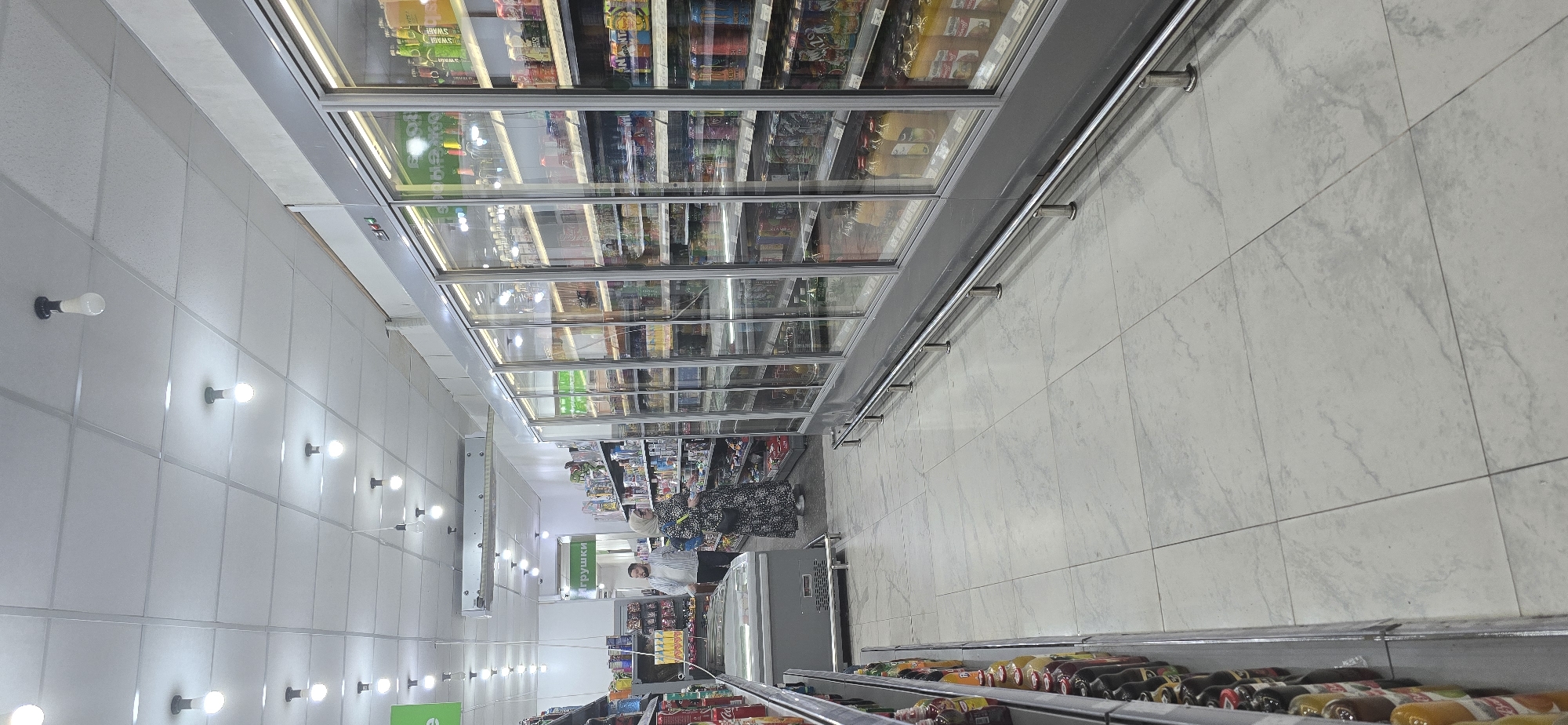 The image depicts the interior of a grocery store, likely in Grozny, Russia. It appears to be a well-lit aisle with refrigerated display cases stocked with various food and beverage products. The floor is tiled with a light-colored, possibly marble or granite, pattern. Fluorescent lights are visible on the ceiling, aligned along the aisle. Shoppers, including at least one woman wearing a headscarf, are present, browsing the items on the shelves. Shelves are stocked with various goods, including beverages.
