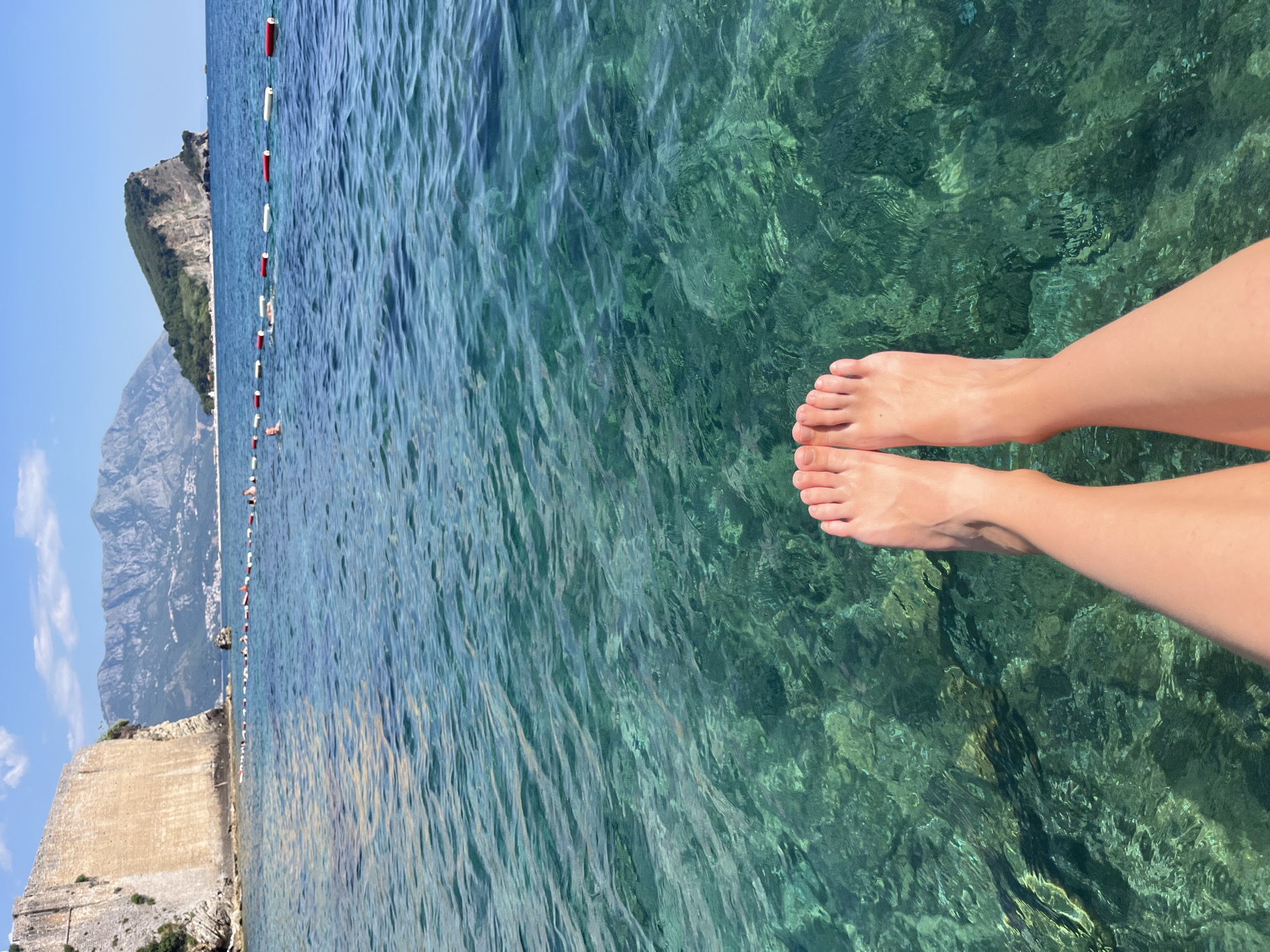 The image depicts a person's legs and feet dangling over clear, turquoise water. The water is calm with gentle ripples, and the sunlight creates shimmering patterns on the sandy bottom visible beneath the surface. In the background, a mountainous coastline with lush green vegetation rises steeply from the sea. A stone wall or fortification runs along the edge of the water, and a line of buoys with red and white markings stretches into the distance, possibly indicating a swimming area or boundary. The sky is a clear, bright blue, suggesting a sunny day. The overall scene evokes a sense of relaxation and a summer vacation. The location is identified as Budva, Montenegro. There is no visible text in the image.