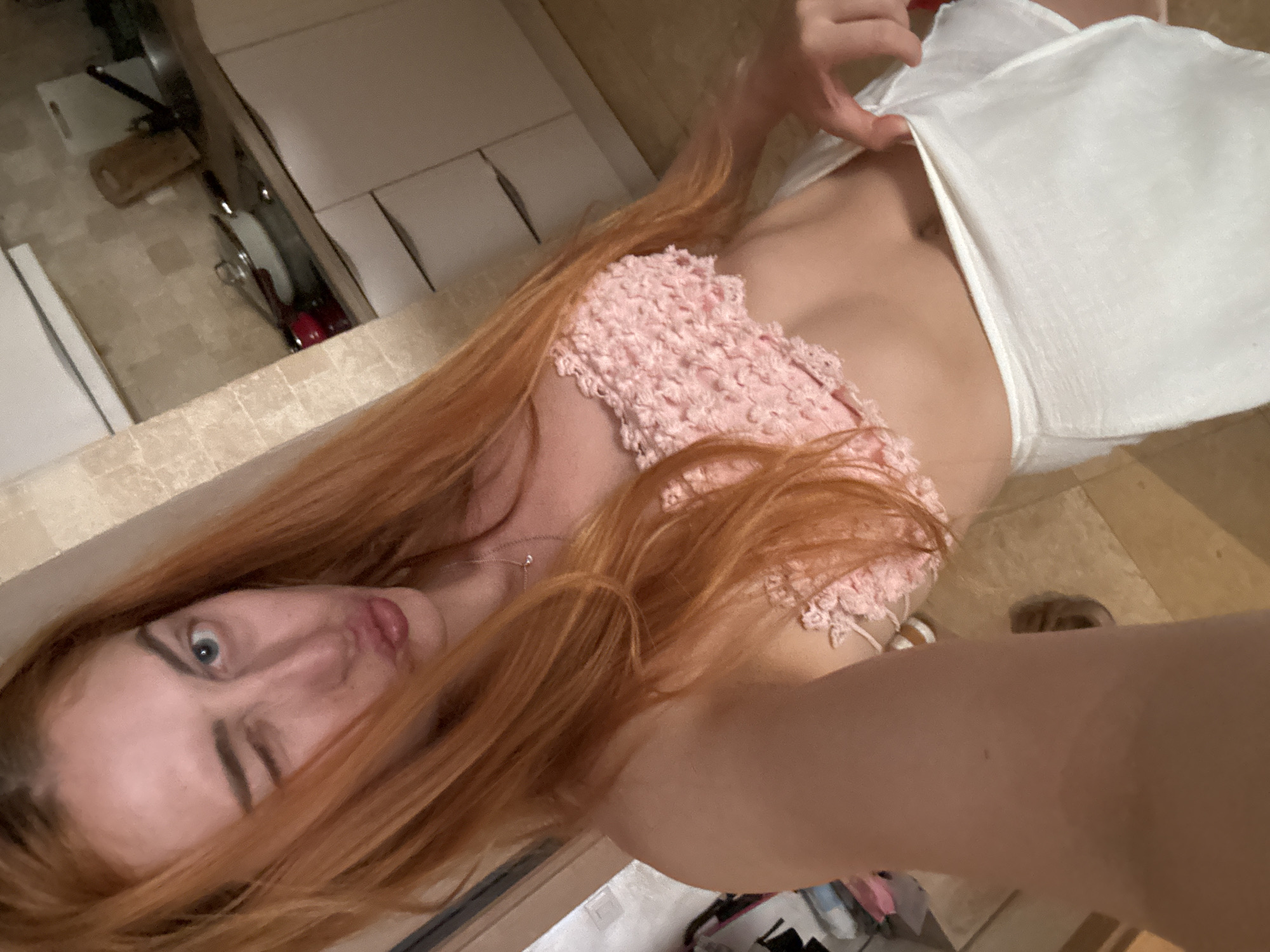 The image is a rotated selfie of a young woman with long, flowing reddish-blonde hair. She lies on her back, looking directly into the camera with one eye partially closed and her lips slightly parted, conveying a neutral to slightly suggestive expression. She is wearing a light pink, textured, possibly crocheted, crop top and a white skirt or wrap. Her right hand is visible, pulling down the waist of her white garment to expose a portion of her hip.
The background, which appears above her head in the rotated view, shows an indoor setting resembling a kitchen or utility area. Light-colored cabinets are visible, and glimpses of kitchenware, such as pots and pans, can be seen stored below. The floor is covered with light-toned tiles. The overall scene suggests an intimate, casual moment captured indoors. While the location context of Budva, Montenegro, was provided, there are no specific visual cues within the image itself to confirm this. The time of day is not discernible due to indoor lighting. No visible text is present in the image.