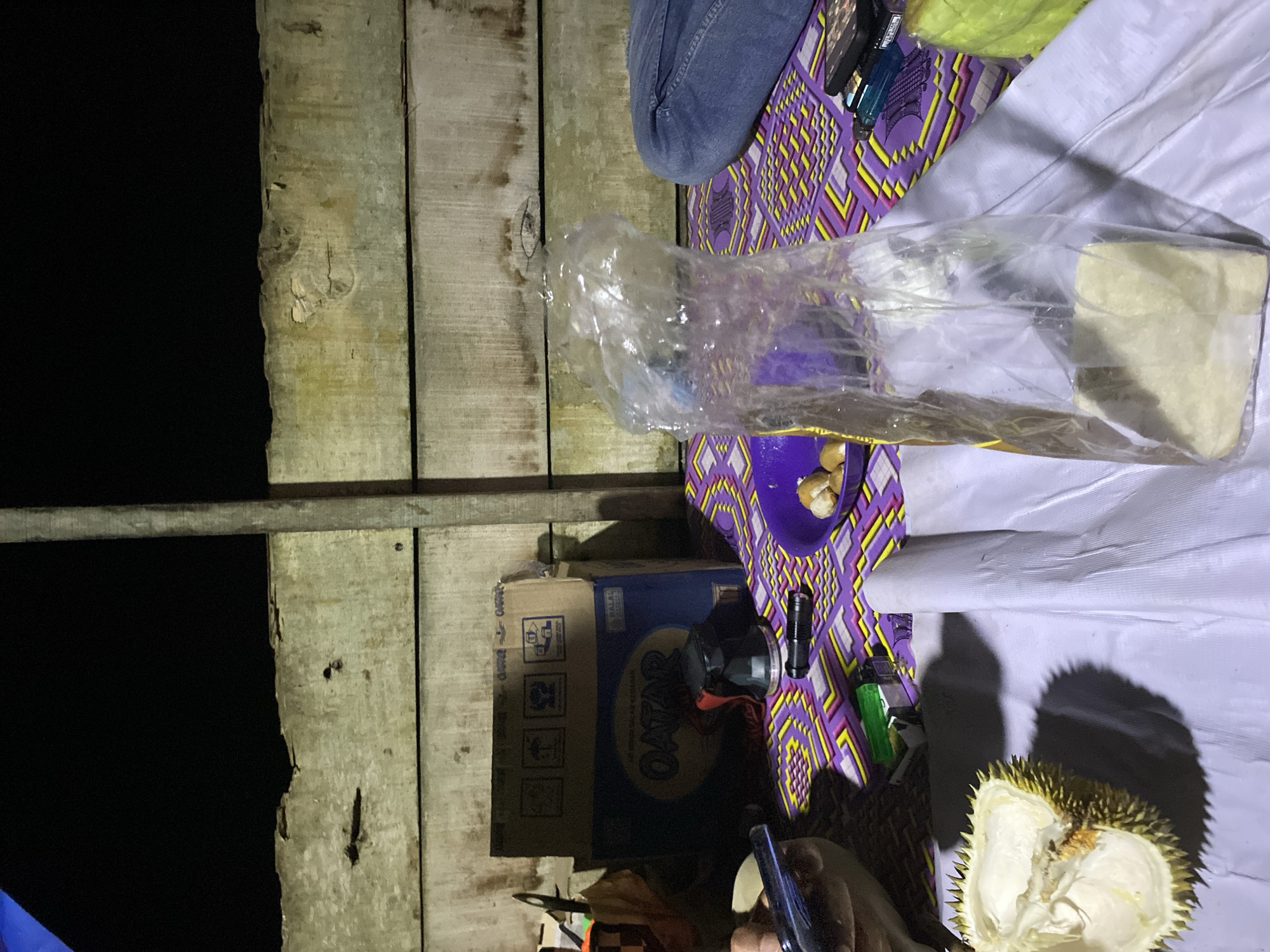 The image depicts an outdoor scene at night, possibly in Lamtui, Indonesia, with a wooden plank wall in the background. In the foreground, a durian fruit is partially opened on a white surface. A purple woven mat with a geometric pattern lies on the ground, holding various items. These include a purple bowl with several small, pale yellow objects, a transparent plastic bag containing a pale yellow substance, a black flashlight, a green lighter, and a smartphone. To the right, a person's legs, clad in blue jeans, are visible, suggesting their presence at the scene. A cardboard box labeled "Qatar" is also present. The overall lighting is dim, with shadows cast by the objects.