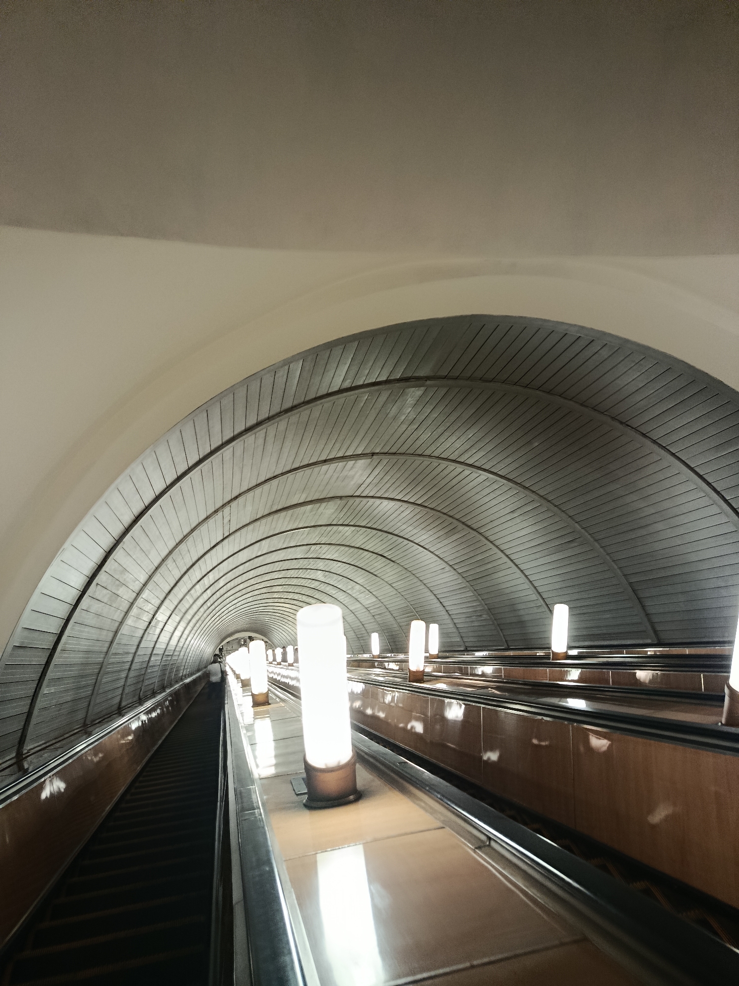The image shows a view from an escalator in a Moscow metro station. The escalator is descending, and the perspective looks down the length of the moving stairs towards a distant person. The station has a distinctive architectural style, with a large, arched ceiling made of segmented metal panels. Along the sides of the escalator, vertical, cylindrical lights are evenly spaced, casting a warm glow. The walls of the escalator are lined with polished brown tiles. The ceiling and walls create a tunnel-like effect, emphasizing the depth and length of the station. The overall impression is one of vastness and perhaps a touch of grandeur, common in many Moscow metro stations. The lighting suggests it is likely daytime, but the interior setting makes specific time of day or weather irrelevant. There is no visible text in the image.