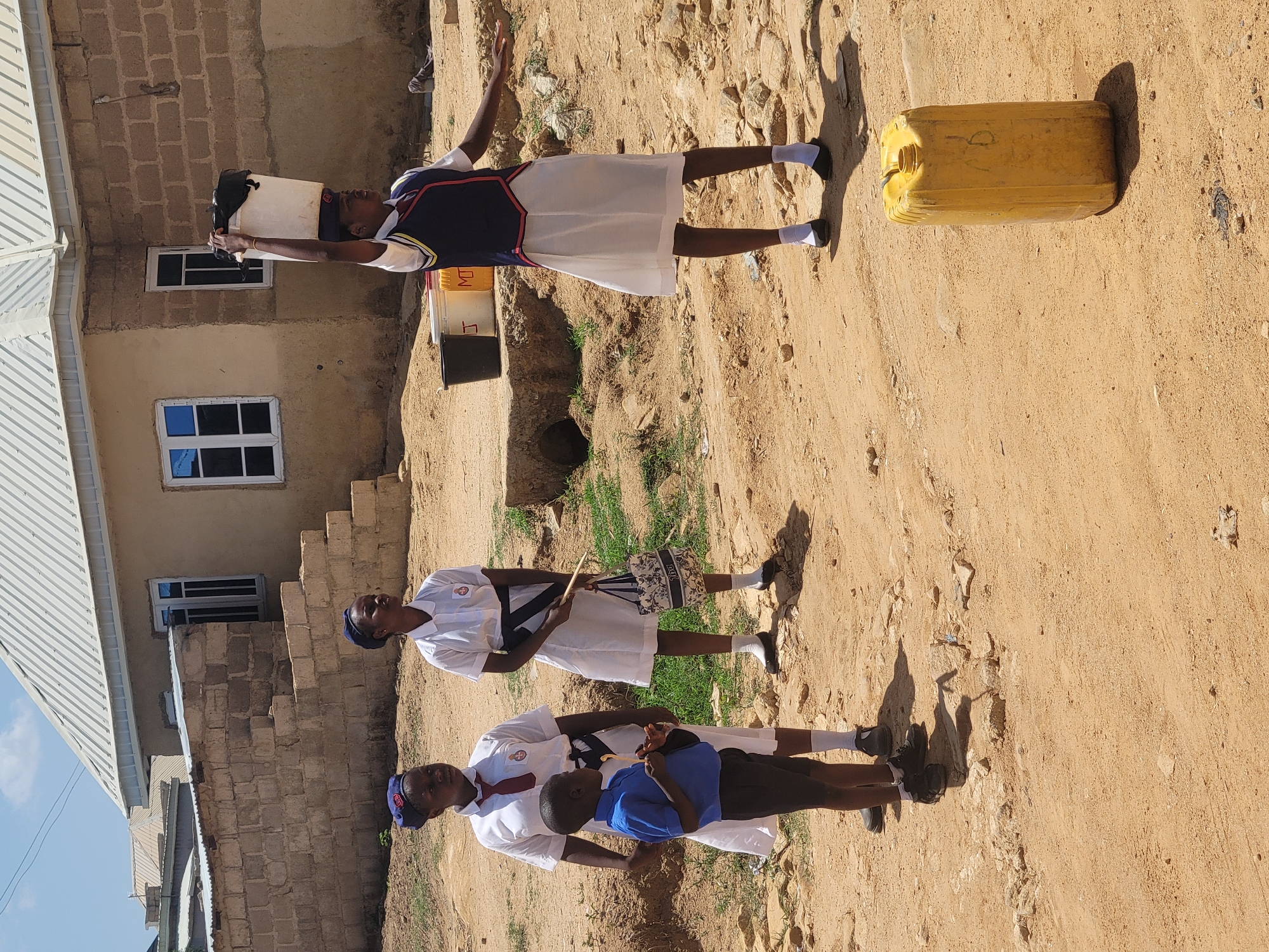 The image depicts three children outdoors, presumably in the city of Jos, Nigeria. They are standing on a dirt or sandy surface in front of a building constructed of brown bricks. The building is multiple stories high and has windows.
One child is wearing a white uniform with a blue vest. She balances a bucket on her head, with her arms raised to maintain balance. Next to her are a bucket, a container, and a yellow jerrycan.
The two other children are wearing white uniforms with blue hats and red ties. One child is holding a bag. The other appears to be helping a younger child dressed in a blue shirt.