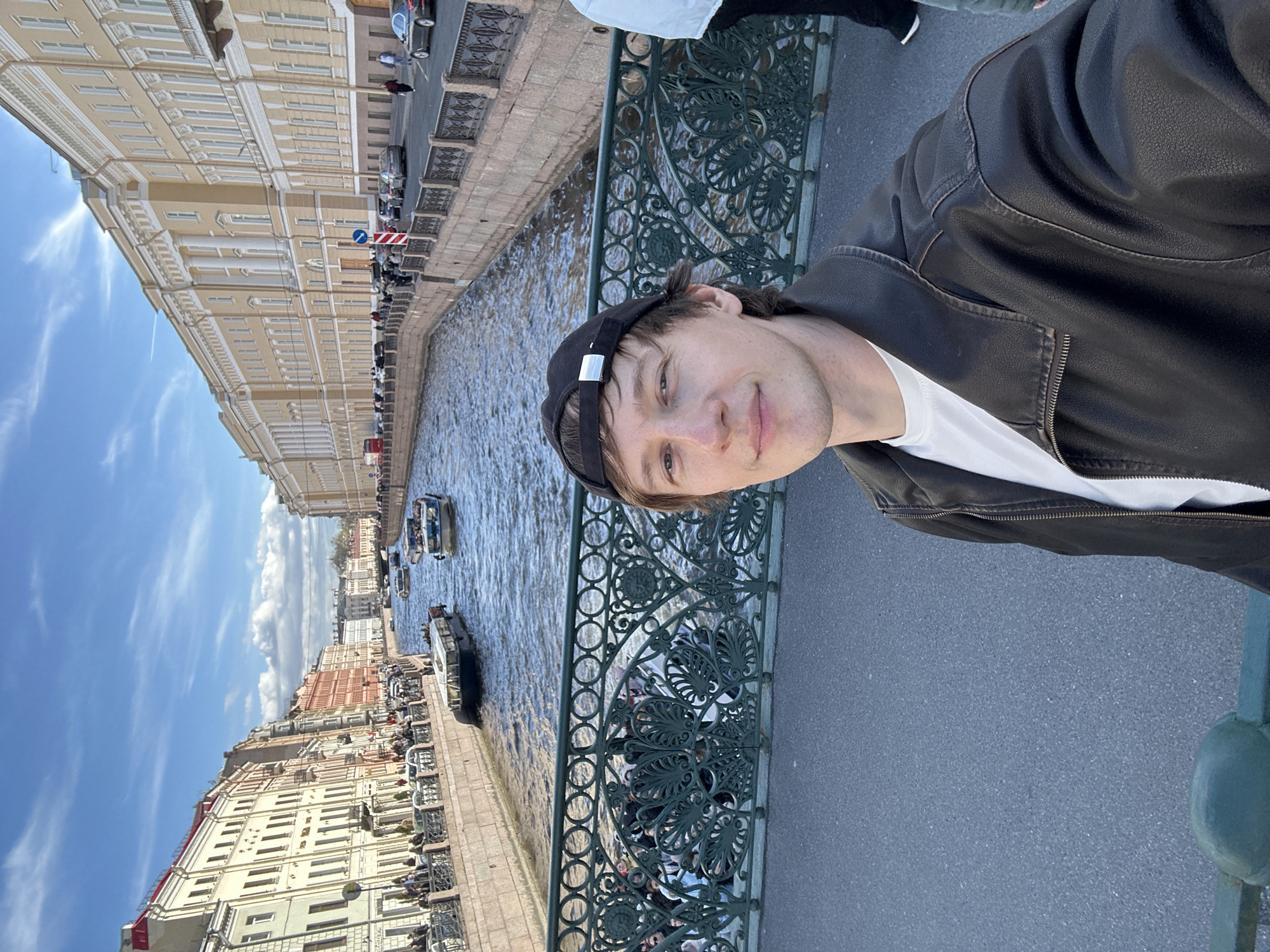 The image is a selfie taken by a man from a bridge overlooking a scenic canal in Saint Petersburg, Russia. The man, positioned on the right side of the frame, is wearing a black baseball cap, a white t-shirt, and a dark brown leather jacket, and has a slight smile. He is standing on a dark grey paved surface next to an ornate dark green metal railing of the bridge, which features intricate circular and foliate patterns.
Behind the man and beneath the bridge, a canal flows with choppy, sun-dappled water. Several boats, likely tour vessels, are visible on the canal, some carrying passengers. Both sides of the canal are lined with grand, multi-story buildings, predominantly in hues of light yellow and beige, characteristic of classical European architecture. People can be seen walking along the embankments, and cars are parked and moving on the street parallel to the canal on the right side of the frame. A blue circular road sign with a red diagonal line, indicating "No Stopping," is visible on the street.
The sky overhead is bright blue with scattered white wispy clouds, suggesting a clear and sunny daytime. The overall scene captures a vibrant day in a historic city.