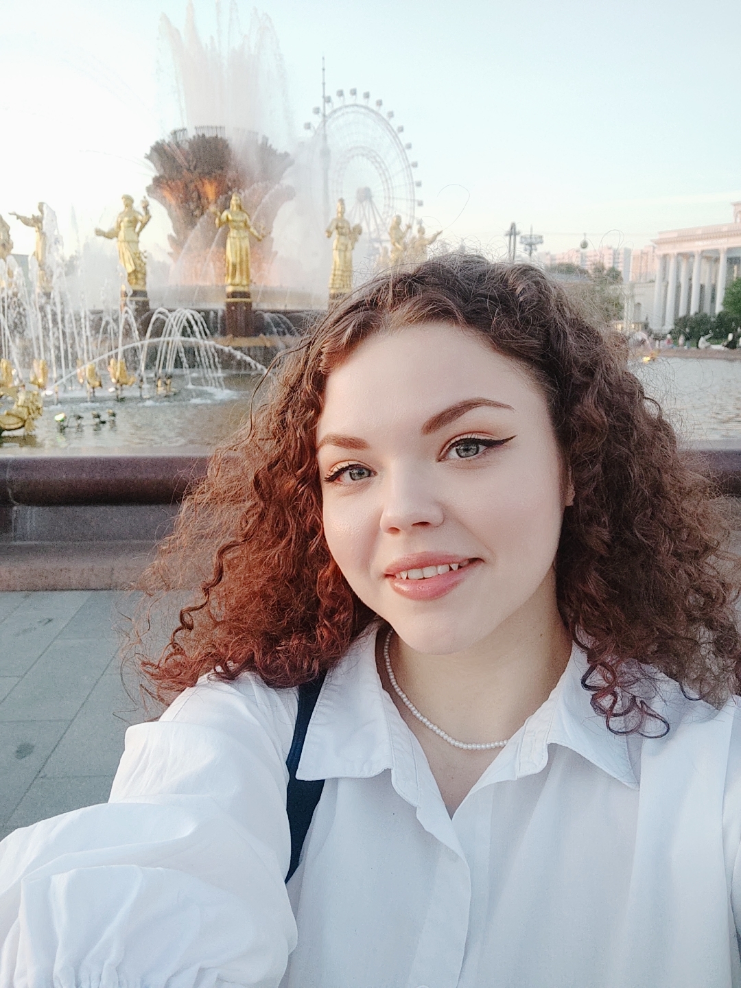 A young woman with curly auburn hair is taking a selfie in front of the Friendship of Nations Fountain at the VDNKh (Exhibition of Achievements of National Economy) in Moscow, Russia. She is wearing a white collared shirt and a delicate pearl necklace. Behind her, the elaborate golden statues of the fountain stand out against the backdrop of the ferris wheel "Moscow-850" and other structures in the park. The water from the fountain is actively spraying, creating a dynamic background. The lighting suggests it's either early morning or late afternoon.