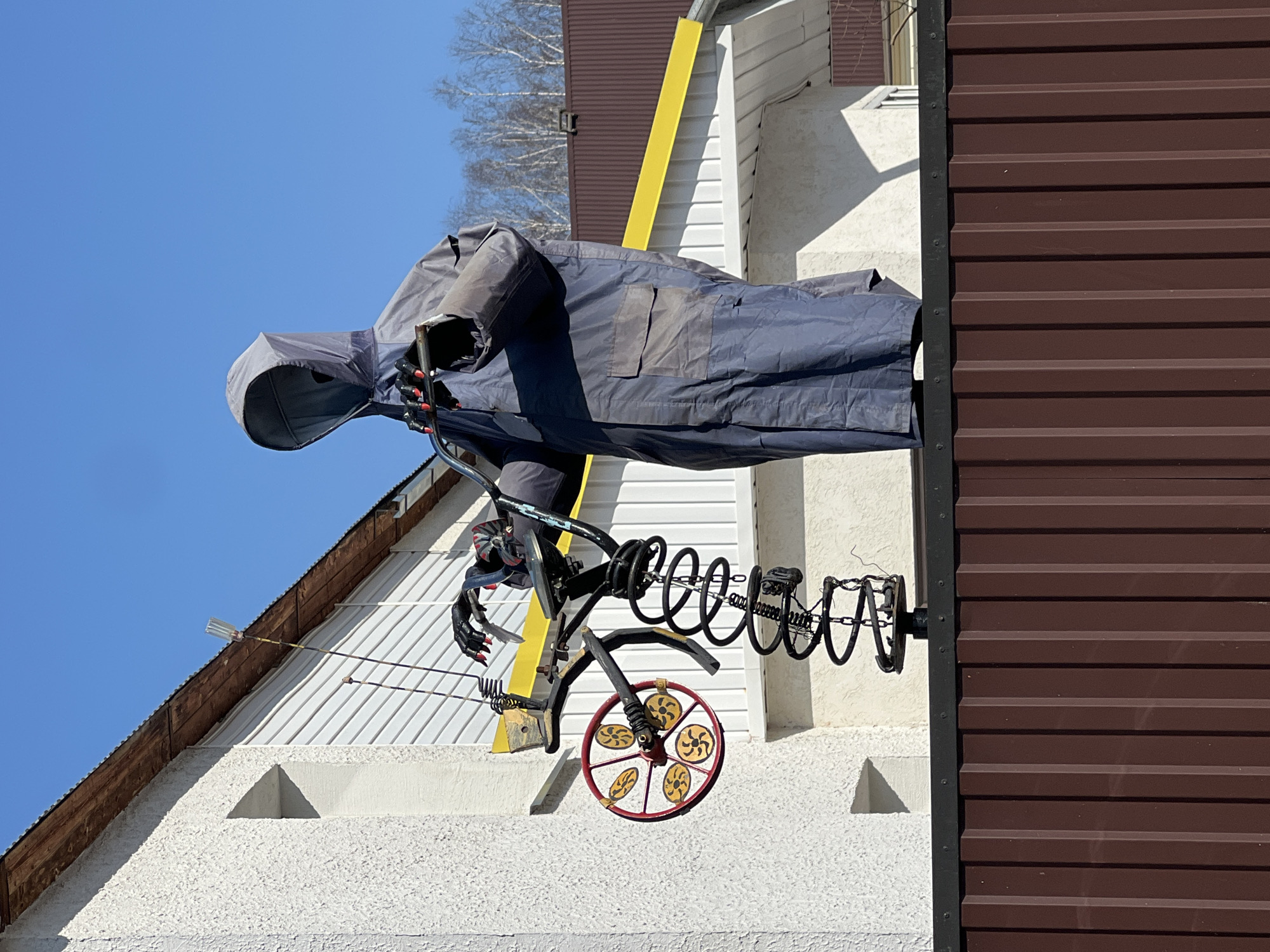 A roadside sculpture depicting a grim reaper on a bicycle is mounted on the side of a building. The grim reaper is fashioned from a dark-colored garment with a hood, obscuring any facial features. The bicycle is constructed from metal, featuring coiled springs and a red-rimmed wheel with gold and red inserts. The building has white stucco walls with triangular indentations, brown corrugated siding, and a yellow vertical accent. The sky is clear and blue. Based on available information, this photograph was taken in Noviy, Russia.
