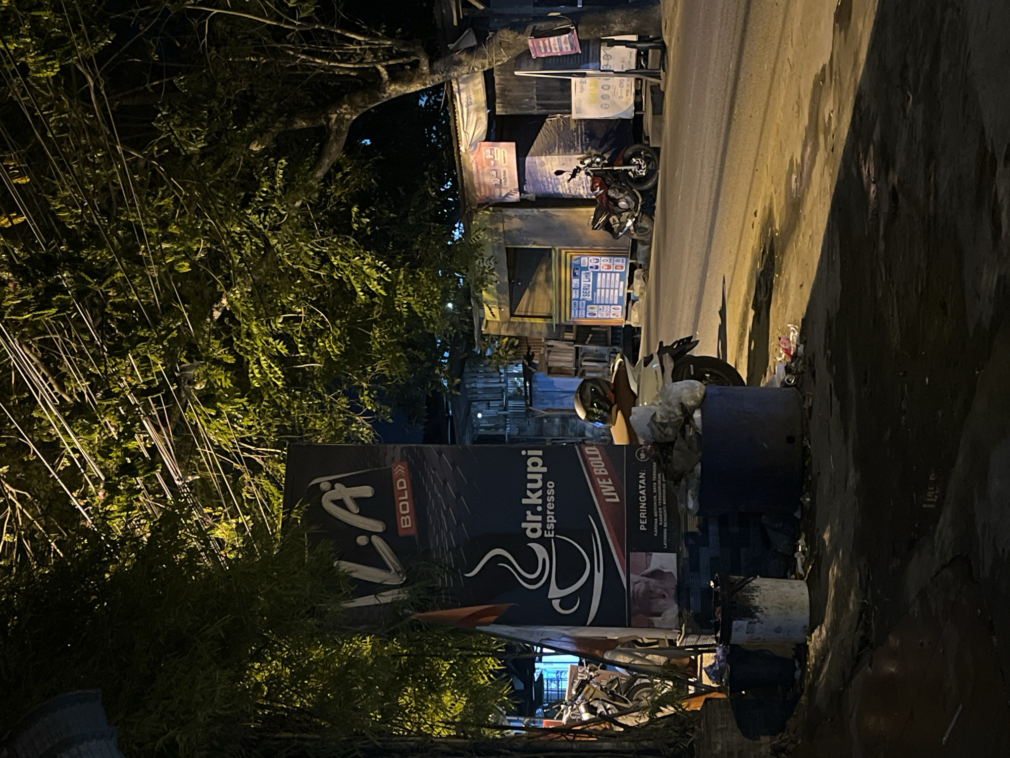 The image depicts a street scene at night in Lhokseumawe, Indonesia, likely near a roadside stall or shop. In the foreground, a large, dark blue sign advertises "LA BOLD" and "dr.kupi Espresso" with a graphic of a coffee cup. Below the coffee cup, there is a warning label with Indonesian text. Two trash cans, one white and paint-splattered and another dark blue, are situated in front of the sign.
The background reveals a street with asphalt pavement. A red motorcycle is parked on the side of the road in front of a building with corrugated metal siding and a banner advertising mobile data packages. To the left of the building, another motorcycle is partially visible. Overhead, numerous electrical wires crisscross through the branches of trees that cast shadows across the scene. The lighting is artificial, emanating from streetlights and nearby establishments, creating a stark contrast between illuminated areas and deep shadows. The overall impression is of a typical, somewhat untidy urban environment at night.