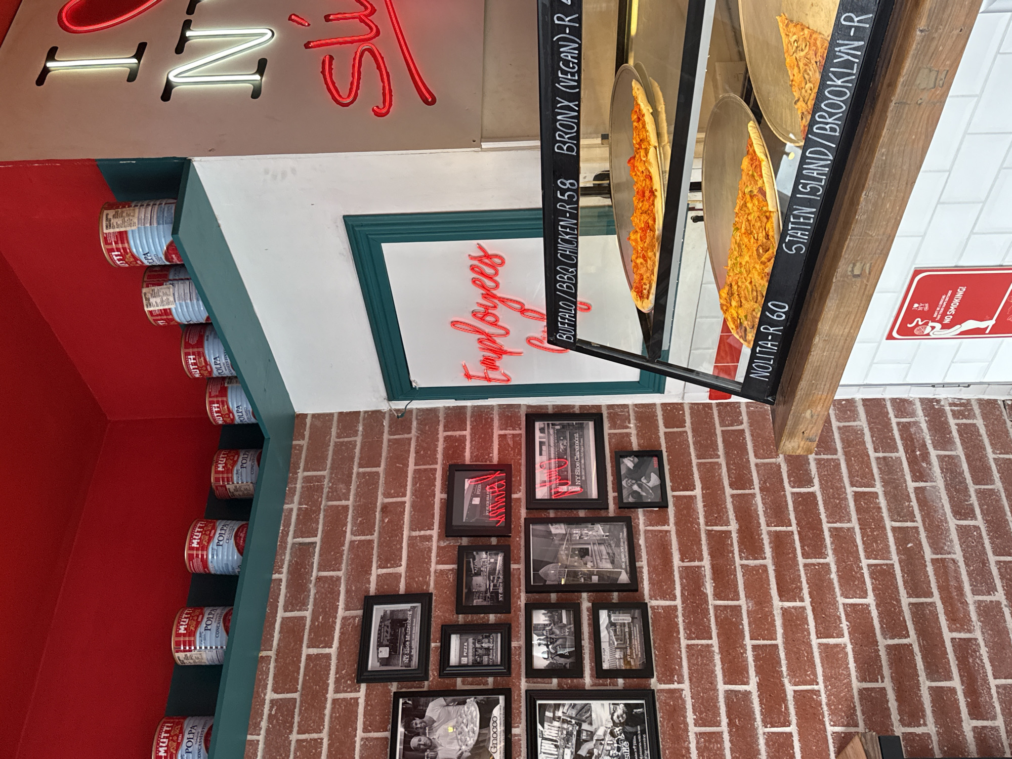The image is taken at a pizza restaurant location, possibly in Cape Town, South Africa. The restaurant is decorated with cans of "MUTTI POLPA" tomato products lined up on a shelf. A green-framed sign with the word "Employees" in red neon hangs on a red brick wall. The brick wall is also decorated with framed black and white photos and images, including photos of "NY Slice Claremont" and "NY Slice Muizenberg". Displayed in a glass case are various pizza slices with labels indicating the flavors: "BUFFALO/BBQ CHICKEN-R58", "BRONX (VEGAN)-R", "NOLITA-R 60", and "STATEN ISLAND/BROOKLYN-R". The price is shown in Rands (R). A "No Smoking" sign is visible on a white tiled wall. A red neon sign is partly visible above the tomato cans, with the letter "N" illuminated.