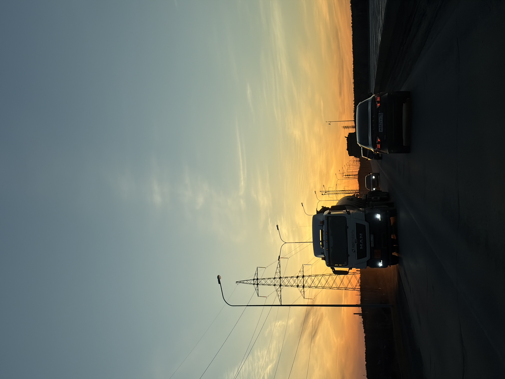 The image shows a road with vehicles at dusk or dawn in Chaikovsky, Russia. A large white MAN truck is traveling towards the viewer, with its headlights illuminated. Behind it, several other vehicles, including a dark car with license plate M447YY, are also visible. The backdrop consists of a gradient sky transitioning from blue to orange near the horizon, indicating either sunrise or sunset. Power lines and streetlights line the roadside. Trees are faintly visible on the horizon.