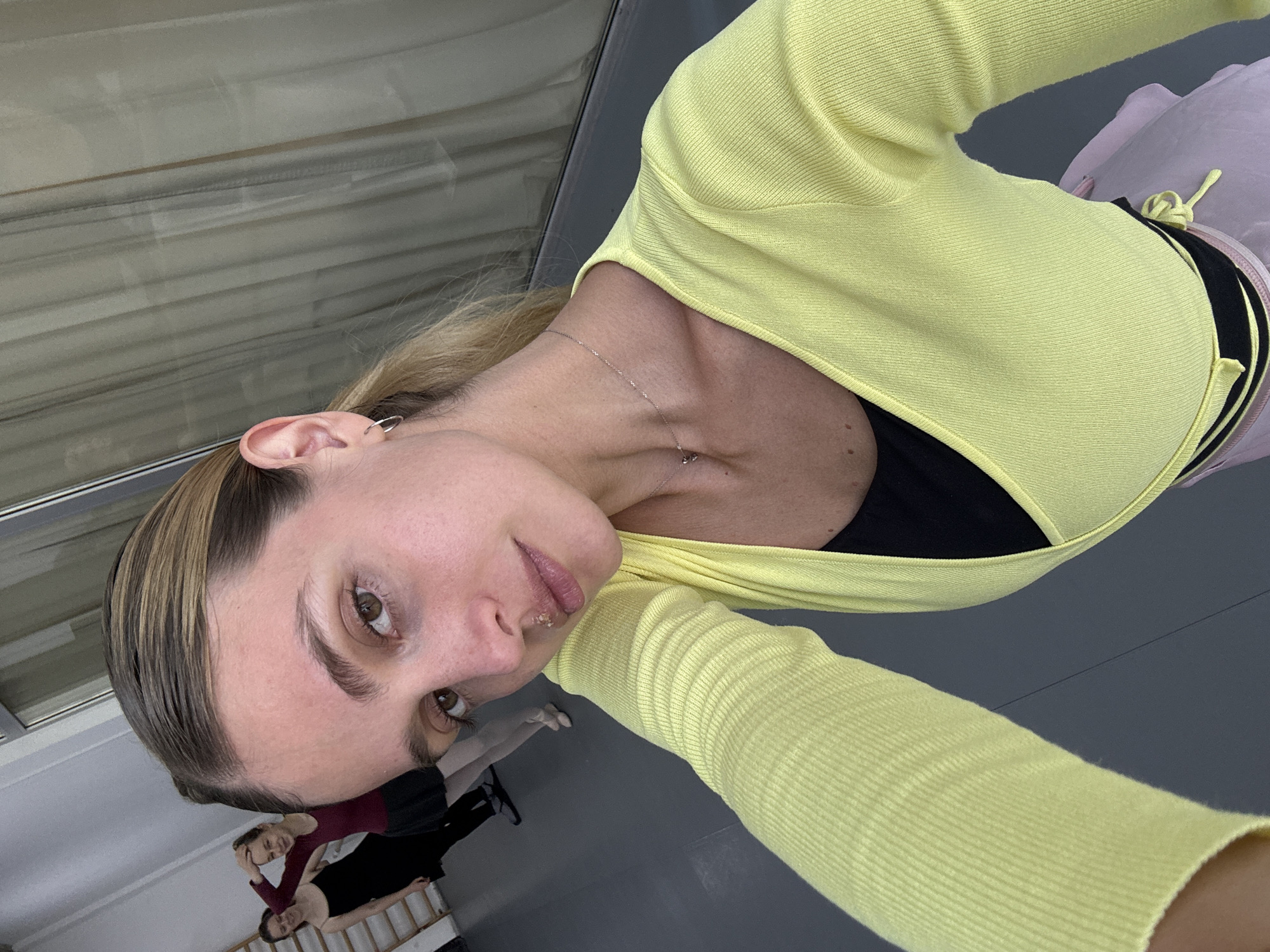 A selfie taken in what appears to be a dance studio. The subject is a woman with blonde hair and brown eyes, wearing a light yellow long-sleeved wrap sweater over a black top, and a light pink skirt tied at the waist. She is facing the camera and looking down. Behind her, two people are visible in the background performing stretches or exercises on the floor. The studio has a gray floor, a wall with horizontal white lines resembling window blinds, and a gray wall. The location is presumably in Tbilisi, Georgia, but there are no specific visual cues in the image to confirm this, and the prompt requested the location regardless.