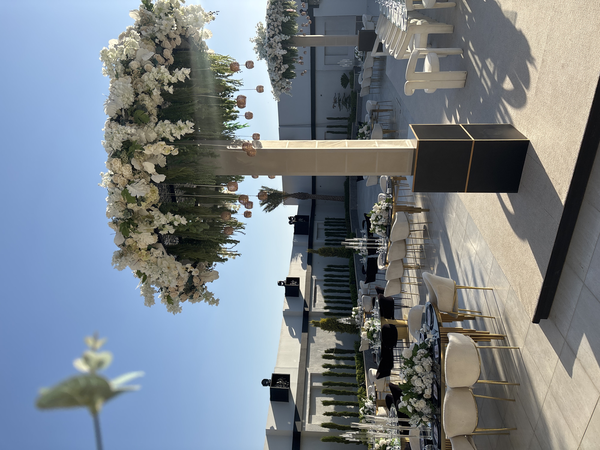 This is an outdoor photograph of a wedding reception setup in Um Rummane, Jordan. The scene features multiple round tables draped in black tablecloths, each adorned with floral centerpieces and glassware. Cream-colored chairs with gold legs surround the tables. In the foreground, a large, ornate floral arrangement hangs from a central pillar, suspended by thin wires and interspersed with decorative globes. Another similar floral structure is visible in the background. The tables are set on a light-colored tiled floor, and a beige carpeted area is present to the right. The background shows a modern building with a flat roof, flanked by rows of tall, thin trees. The sky is clear and blue. There are no people visible in the frame.