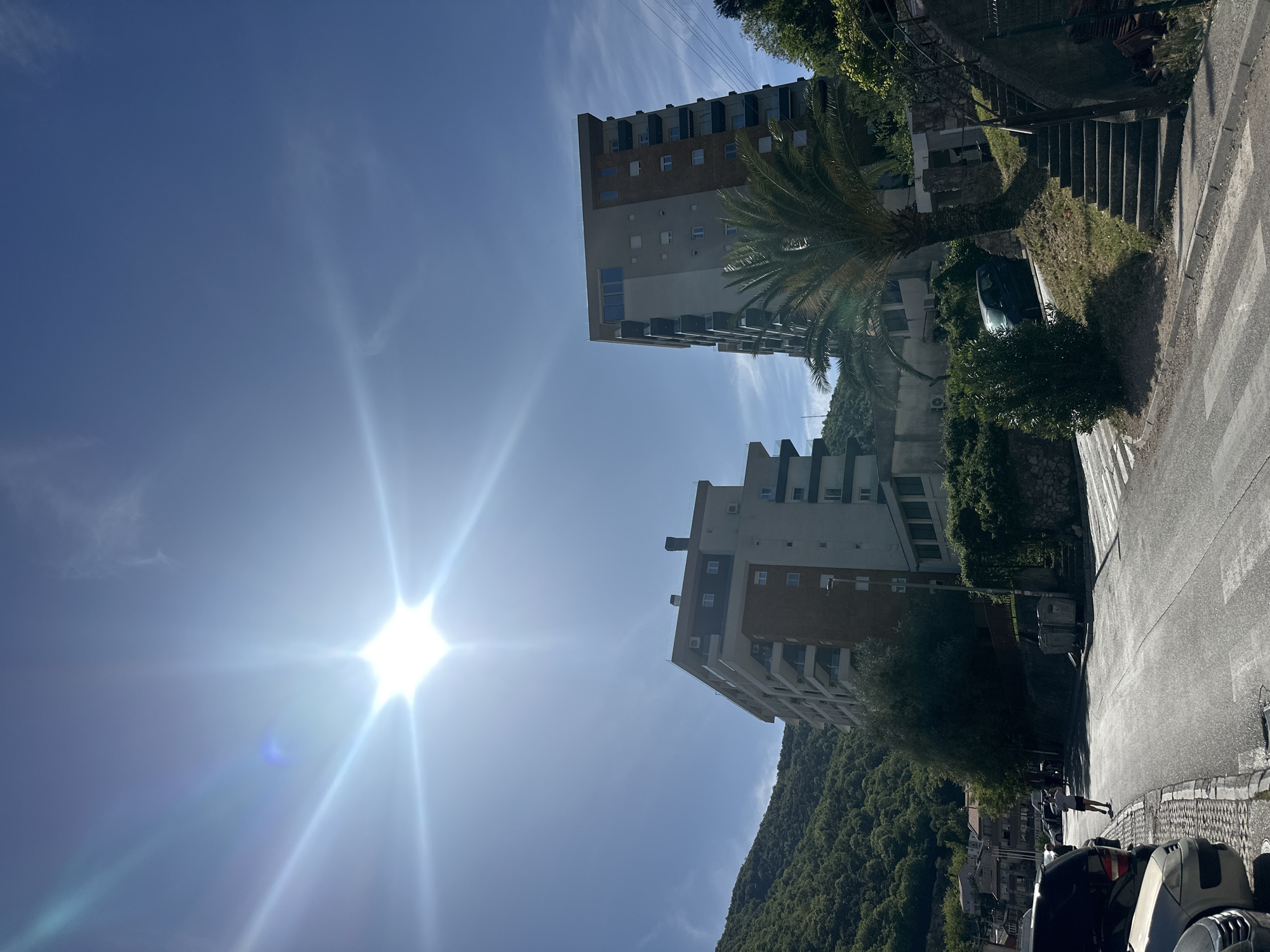 This media file captures a bright, sunny daytime scene in what appears to be a residential or mixed-use area, identified as Budva, Montenegro. The image is taken from a street level, looking upwards, with the sun prominently visible in the sky.
In the foreground, a paved street with a zebra crossing is visible on the right. Several cars are parked along the side of the road, including a dark-colored vehicle. A person wearing a white t-shirt and dark shorts is walking on the sidewalk. To the left of the road, an embankment covered in green grass and lush vegetation, including palm trees, slopes upwards, featuring a set of concrete stairs.
The middle ground is dominated by two modern-looking, multi-story apartment buildings. The one on the left is taller, with multiple balconies and a facade combining light and brown sections. The building on the right is slightly shorter and also has balconies. Green trees and other vegetation surround the buildings. Power lines are faintly visible against the sky near the top left.
In the background, a large, verdant mountain covered in dense trees rises, indicating the mountainous landscape of the region. The sky is a clear, bright blue with minimal, wispy clouds. The sun is positioned in the upper left portion of the frame, creating a strong, star-like lens flare that radiates across a significant part of the sky, indicating direct sunlight.
The activity primarily involves a person walking and cars parked, suggesting everyday urban life. The time of day is clearly daytime, characterized by bright sunlight and clear skies, indicating good weather. No discernible text is visible on signs or vehicles. The scene depicts a typical Mediterranean or Adriatic town environment with a blend of architecture, natural elements, and urban infrastructure.