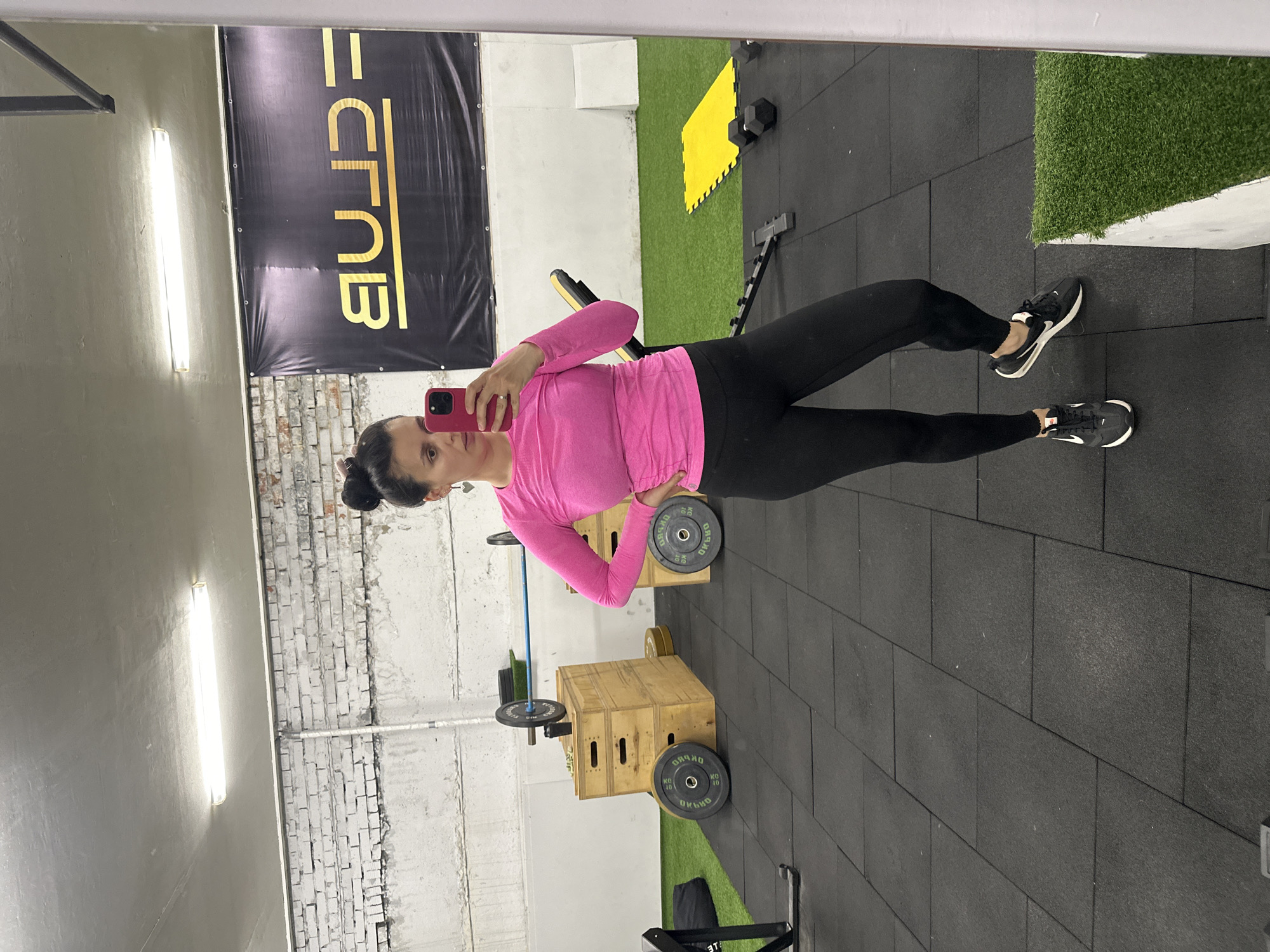 The image is a selfie taken in a gym. The person in the image is a woman with dark hair in a bun, wearing a pink long-sleeved shirt and black leggings. She is holding a phone in her left hand and looking at the camera. The background of the image shows gym equipment, including weights, plyometric boxes, and a weight bench. There is a banner on the wall with yellow text, likely the gym's name. Based on the banner text, the gym could be identified as a fitness club located in Chaikovsky, Russia. The flooring is black rubber. The walls appear to be painted white or grey, with exposed brickwork visible in some areas. Artificial grass is also present in a corner of the gym.