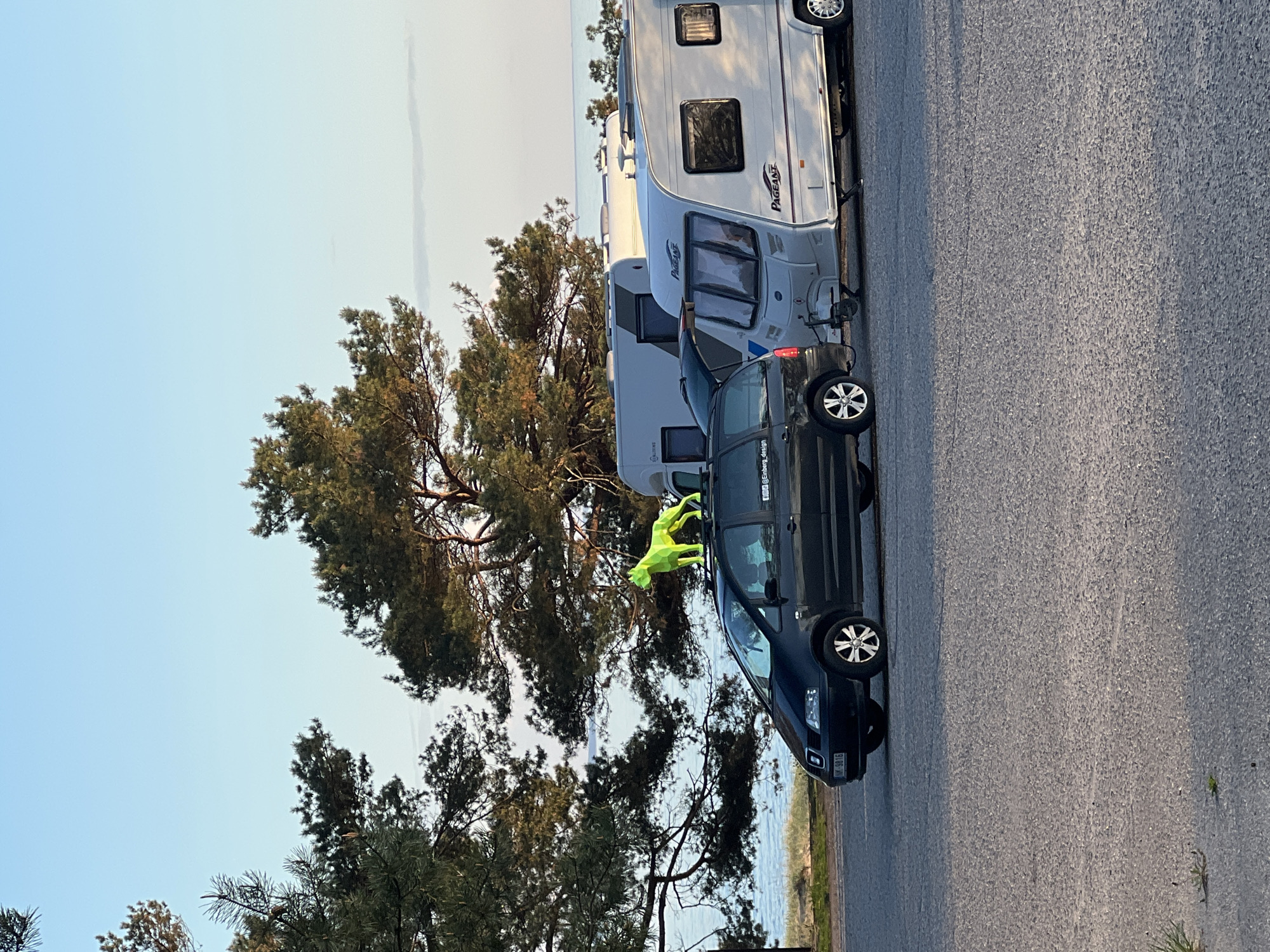 The image shows a partially overcast outdoor scene, likely a parking area near the sea. A black car is parked adjacent to two white trailers, one behind the other. A neon green figure is perched atop the car, appearing to be a decorative or promotional item. The background includes a large tree, the Baltic Sea, and the sky. The scene is located in Saulkrasti, Latvia.
