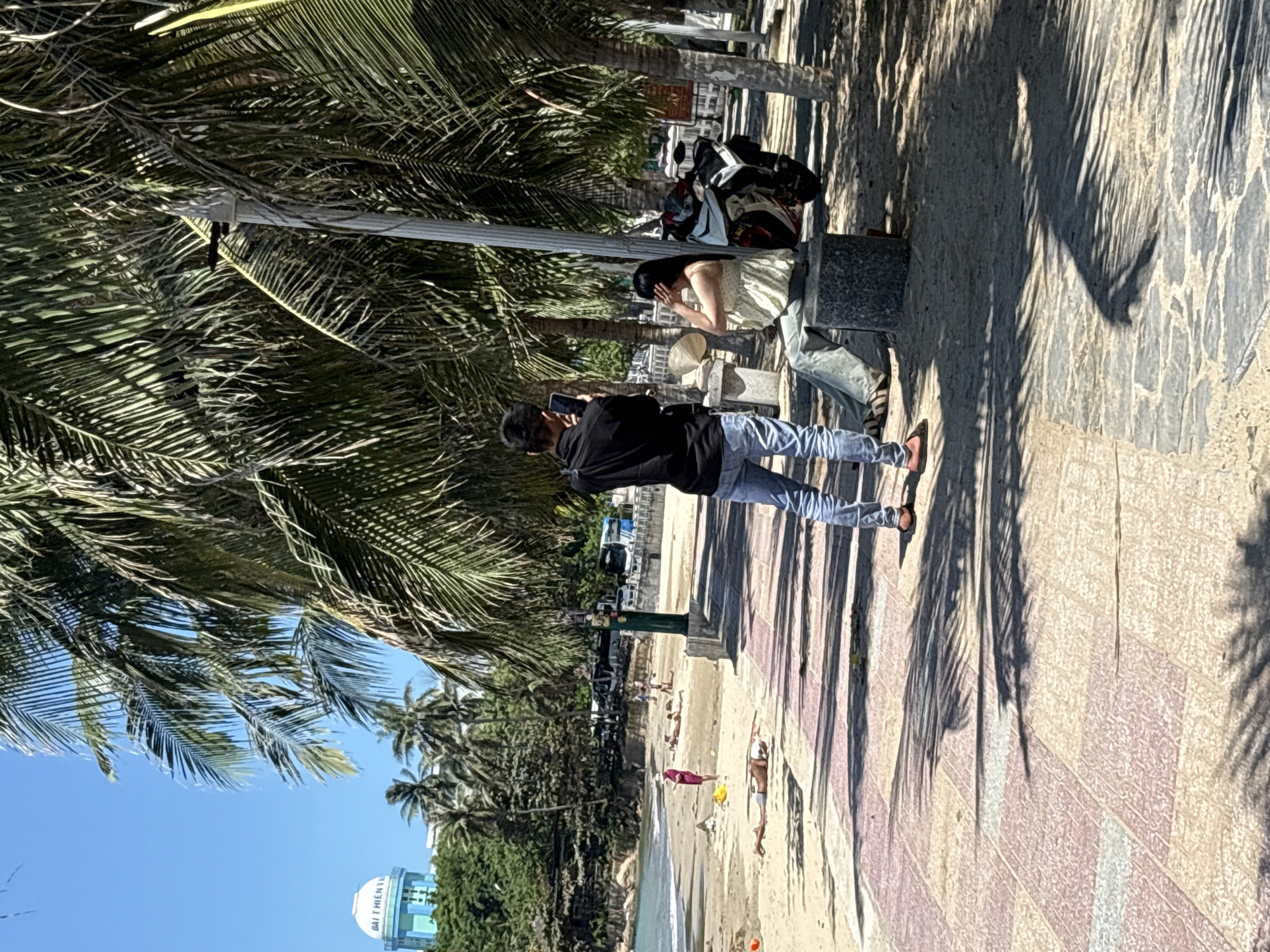 A sunny day in Bac Nha Trang Ward, Vietnam, shows a paved area next to a beach with palm trees. A man in a black jacket and blue jeans is standing on the paved area, holding a phone as if taking a picture. Near him, a woman in a light-colored dress is sitting on a concrete block. Behind them, a parked motorcycle is visible. In the background, the beach is populated with people, some sunbathing. Further off, trees and a water tower are visible. The scene is cast with shadows from the palm trees.
