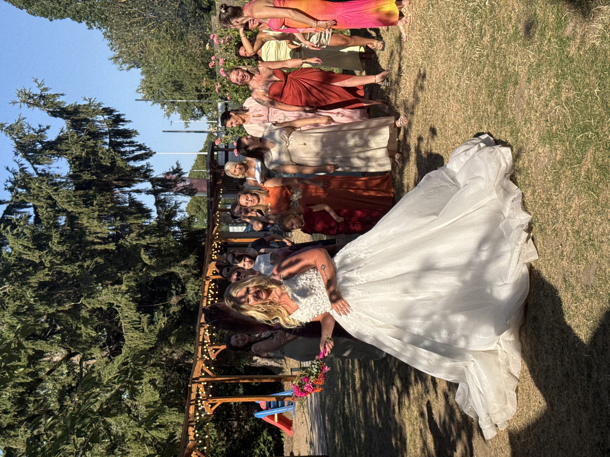 The image depicts an outdoor wedding scene, possibly in a garden or park setting, with a bride in a white gown at the forefront, holding a bouquet. She is facing towards the viewer and appears to be smiling. Behind her, a group of women, presumably bridesmaids or guests, are gathered, some in colorful dresses. The environment is sunny with visible trees and greenery. There is a wooden structure with lights in the background, and a children's slide is visible to the left. The ground is covered with dry grass. The image does not contain any information about the city of Hem, France.