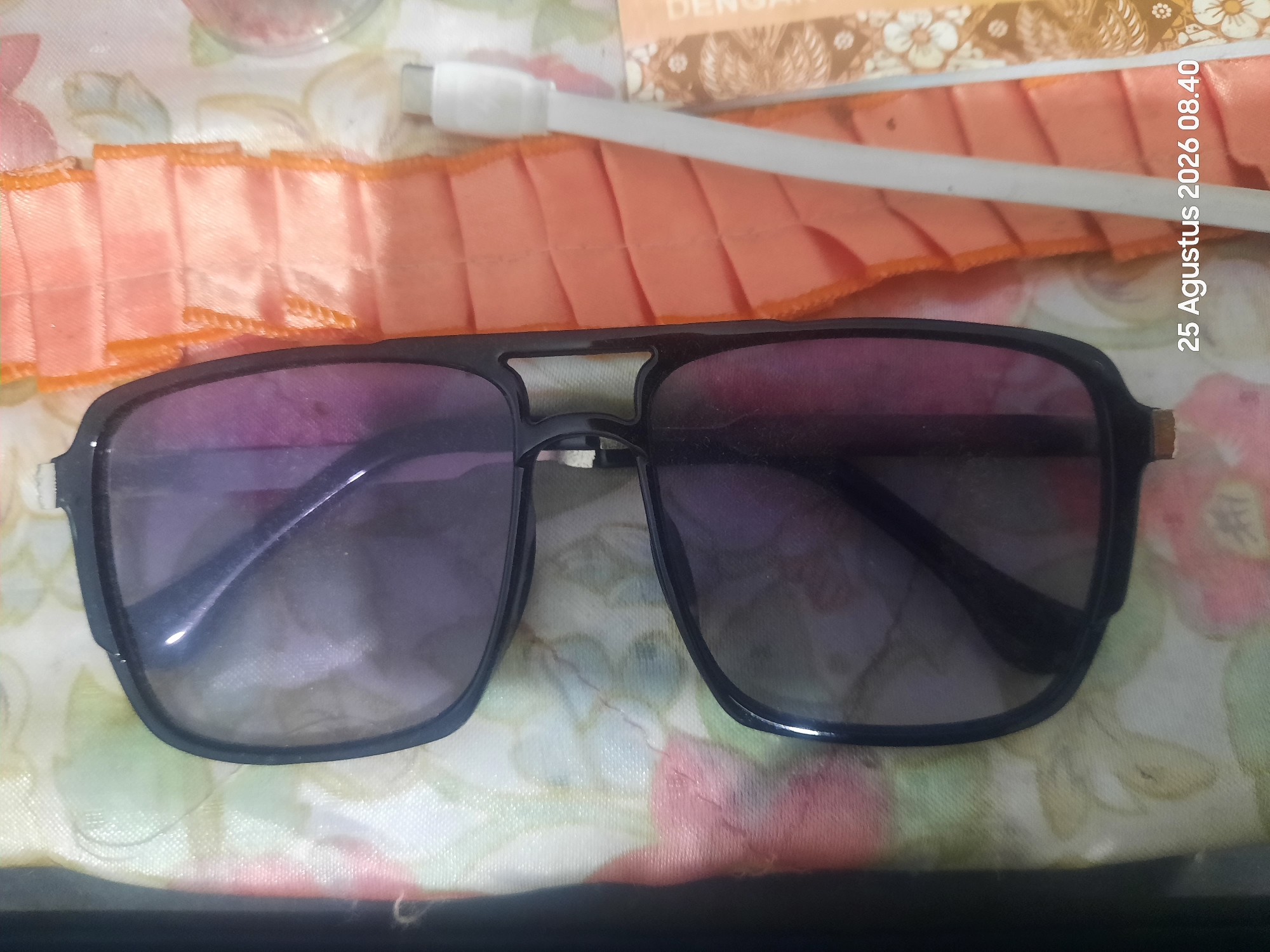 A pair of dark-rimmed sunglasses with purple lenses lies on a patterned fabric surface. To the right of the sunglasses, a section of orange pleated fabric is visible, next to a white USB cable. In the bottom right corner, a date and time stamp reads "25 Agustus 2026 08.40". The scene appears to be indoors, with no discernible features of the city of Sukokerto, Indonesia.