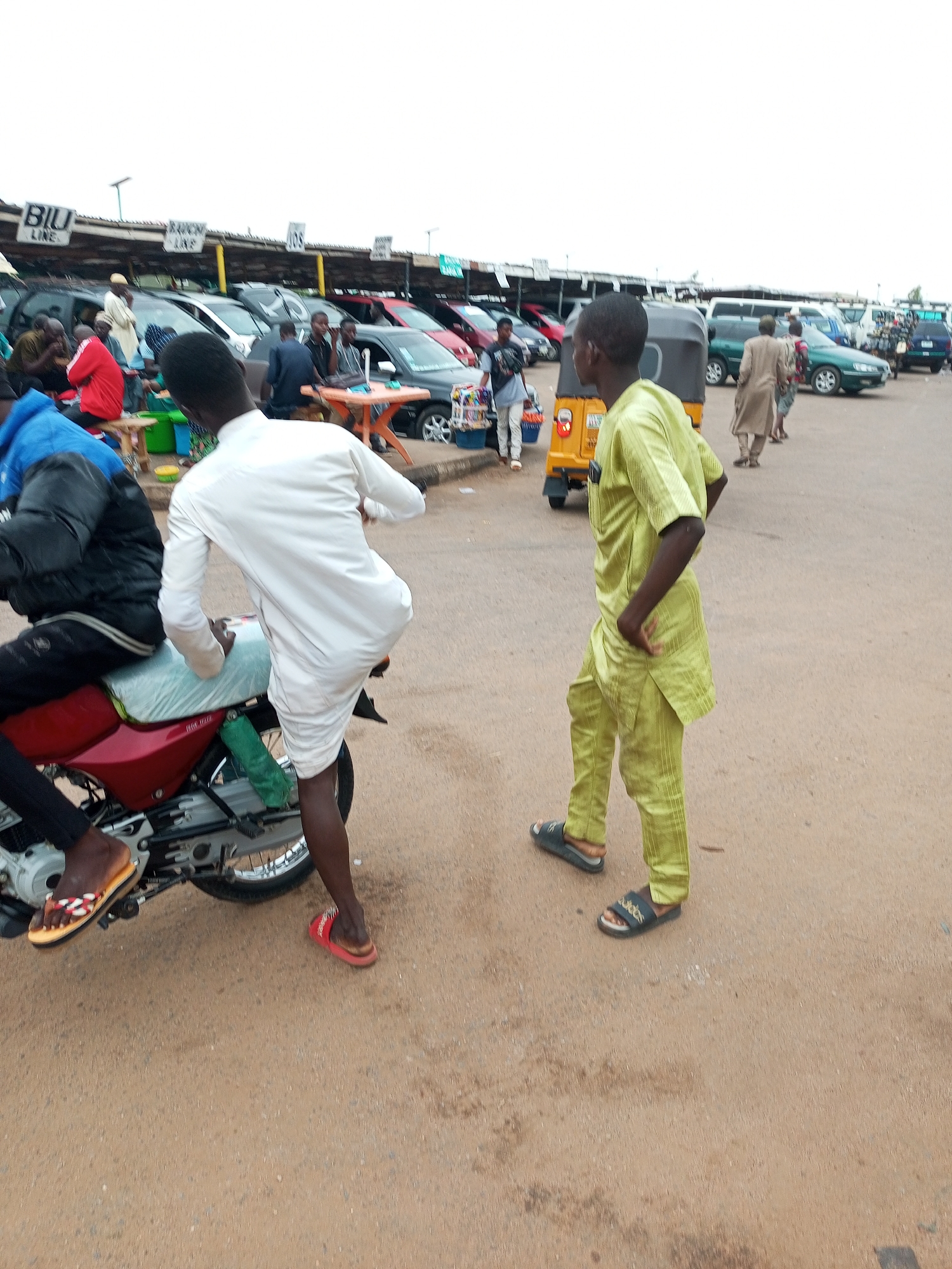 In Gombe, Nigeria, a motorcyclist wearing a white robe and red sandals is seated on a red motorcycle. He is facing left, with his legs straddling the motorcycle. A person wearing a green tunic and pants, with black sandals, stands to the right of the motorcyclist, facing left. In the background, several vehicles, including cars and a tricycle, are parked in a lot under a covered structure. People are seen milling about in the background. The ground is covered in sand and gravel.