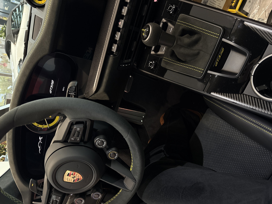 The image provides an internal view of a black Porsche 911 GT3 RS, situated within what appears to be a Porsche dealership in Regensburg, Germany. The perspective is from the driver's seat, looking towards the dashboard and center console.
The steering wheel is trimmed in black Alcantara with yellow contrast stitching and features the Porsche crest centrally. Control buttons, including "DRIVE MODE" and "P-TCC" (likely Porsche Traction Control), are visible on the lower spokes.
The digital dashboard display indicates "GT3 RS" and a temperature of "24.5°C," along with a white silhouette of a car. A yellow-accented analog tachometer is partially visible to the right. The passenger side of the dashboard is trimmed with a carbon fiber pattern.
The center console is finished in glossy black, featuring carbon fiber accents along the edges of the driver's side. A manual gear shifter, covered in black Alcantara with yellow stitching and displaying a yellow gear pattern (1 through 6 and R), is prominent. The "GT3 RS" designation is embossed on the console surround below the shifter. A single cupholder is located towards the front right of the console.
A person's left leg, clad in black pants, occupies the driver's seat, which features a perforated pattern and yellow stitching.
Through a large glass partition in the background, another white convertible Porsche vehicle is partially visible, indicating a showroom environment. A blurred gold spherical object is also discernible in the distant background.
