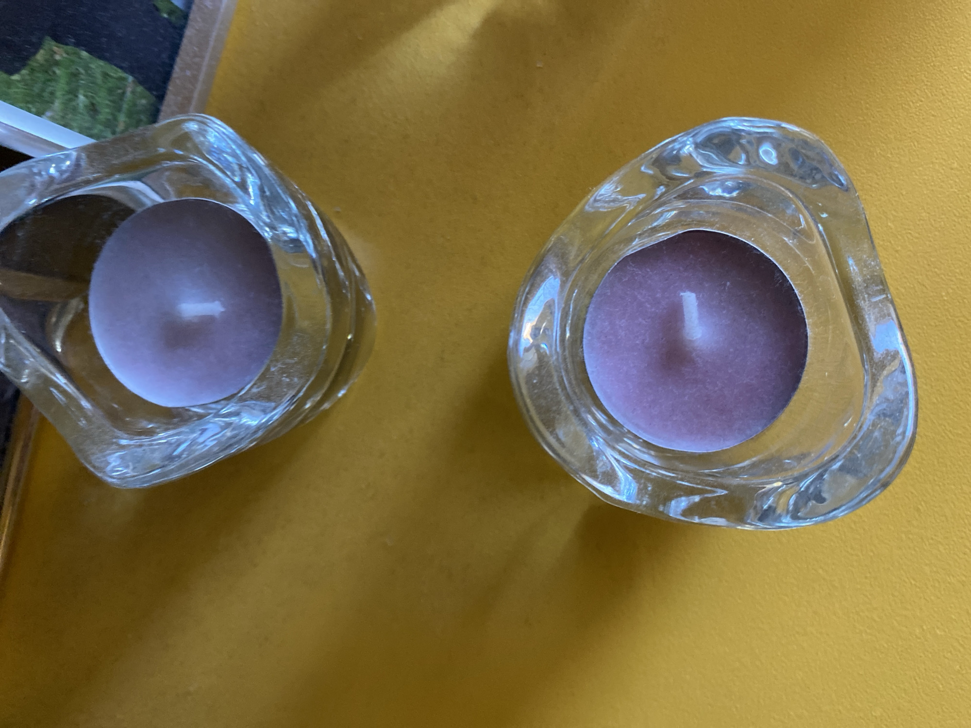 Two purple votive candles in uniquely shaped clear glass holders are placed on a textured golden surface. The candle in the upper holder is centered, and the candle in the lower holder is slightly off-center. The surface reflects light, creating shadows and highlights. A portion of a framed picture with a green grassy background and a dark silhouette is visible in the upper right corner of the image. The scene appears to be an indoor setting, possibly a table or shelf, in a home or business located in Varna, Bulgaria.