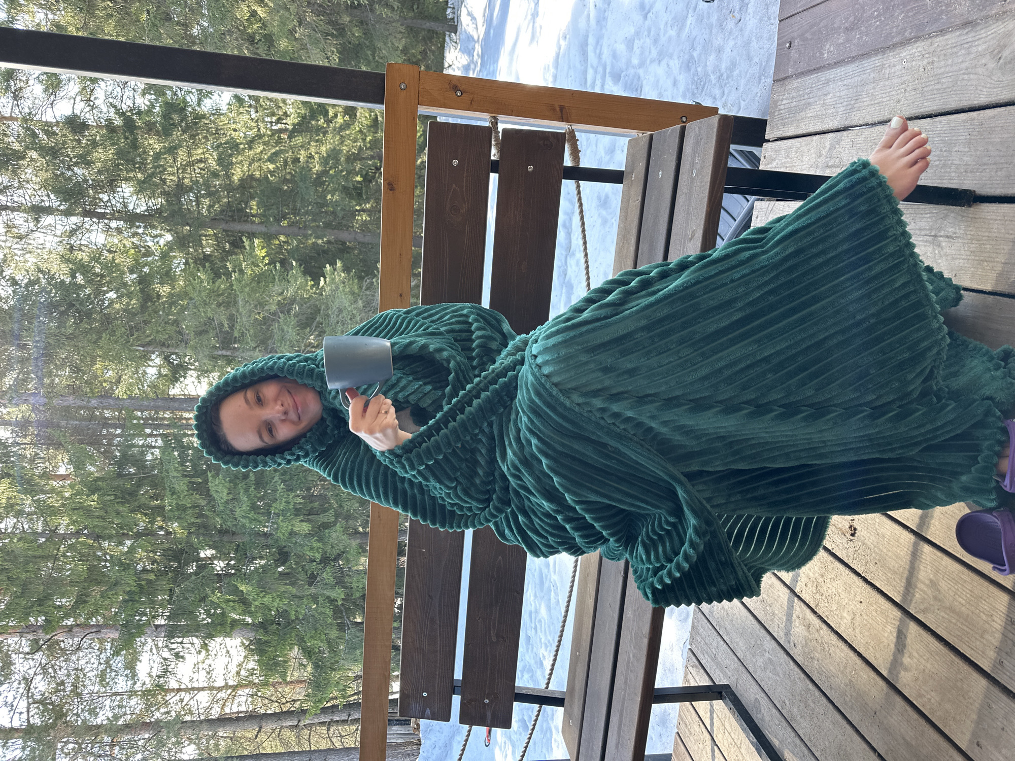 An adult female is depicted seated on a wooden bench located on an outdoor deck in Прикамский, Russia. She is wrapped in a dark green, ribbed, hooded blanket, holding a grey mug with her right hand; a wedding band is visible on her left ring finger. Her bare right foot, adorned with white nail polish, rests on the wooden plank flooring, adjacent to a purple clog shoe. The subject is looking towards the camera with a slight smile. The background consists of a dense forest of tall evergreen trees, with snow visible on the ground beyond the wooden deck, indicating a winter or early spring setting under bright sunlight. The deck features a wooden bench with dark brown slats and a lighter wooden frame, along with a rope railing and a vertical dark support beam on the right side.