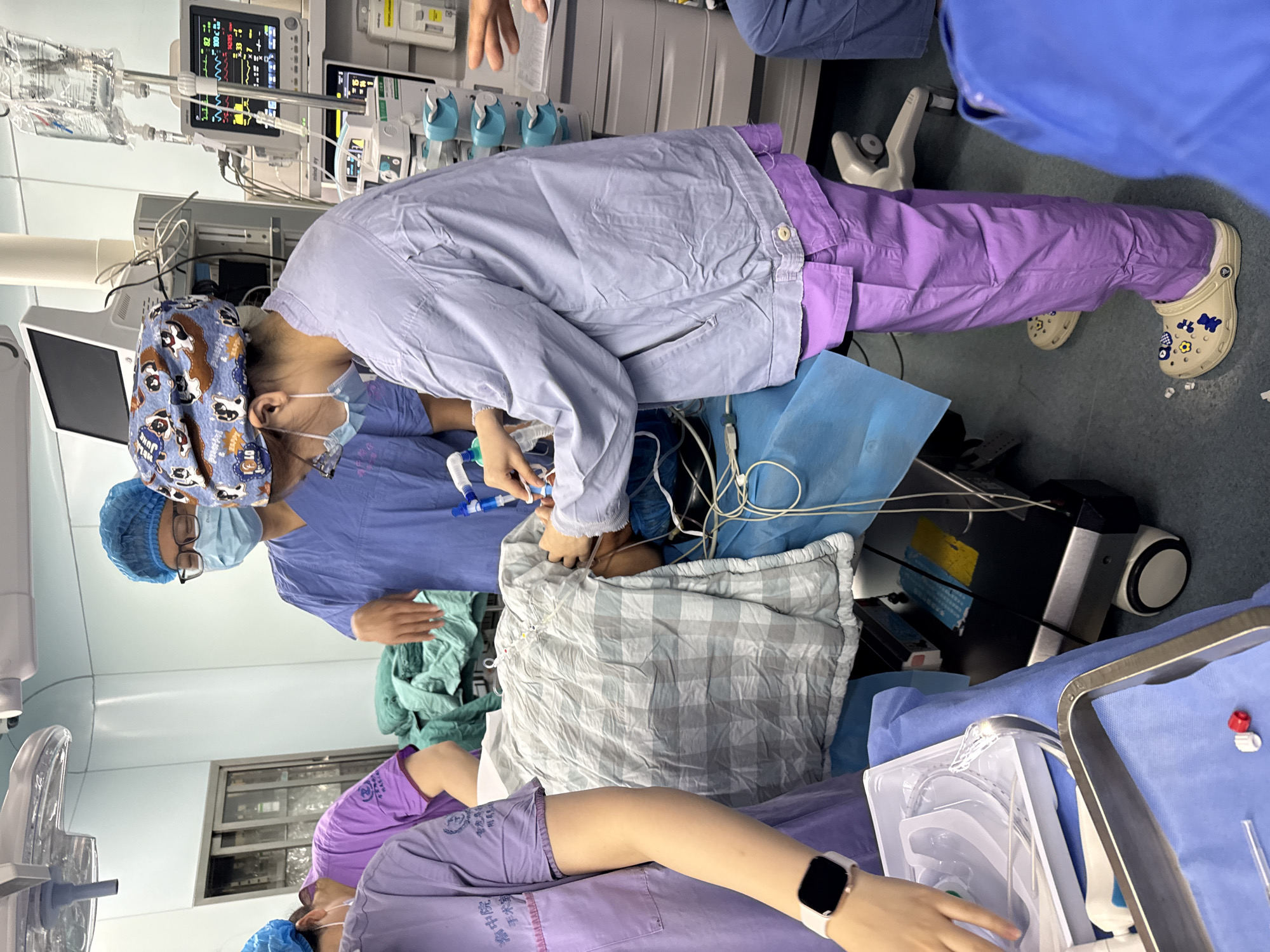 A surgical operation is underway in what appears to be an operating room. Several medical professionals in surgical attire are attending to a patient lying on a table covered with blue surgical drapes. One individual, wearing a surgical cap with a cartoon pattern, is leaning over the patient, possibly performing a procedure. The patient is covered with a gray and white checkered blanket.
Medical equipment, including a monitor displaying vital signs, is visible in the background. Medical instruments are on a tray next to the patient. Another person, wearing purple surgical scrubs and a watch, is within the frame. One person is wearing Crocs shoes with decorations.
The location, Yuzhong District, China, is not discernible from the visual information provided.