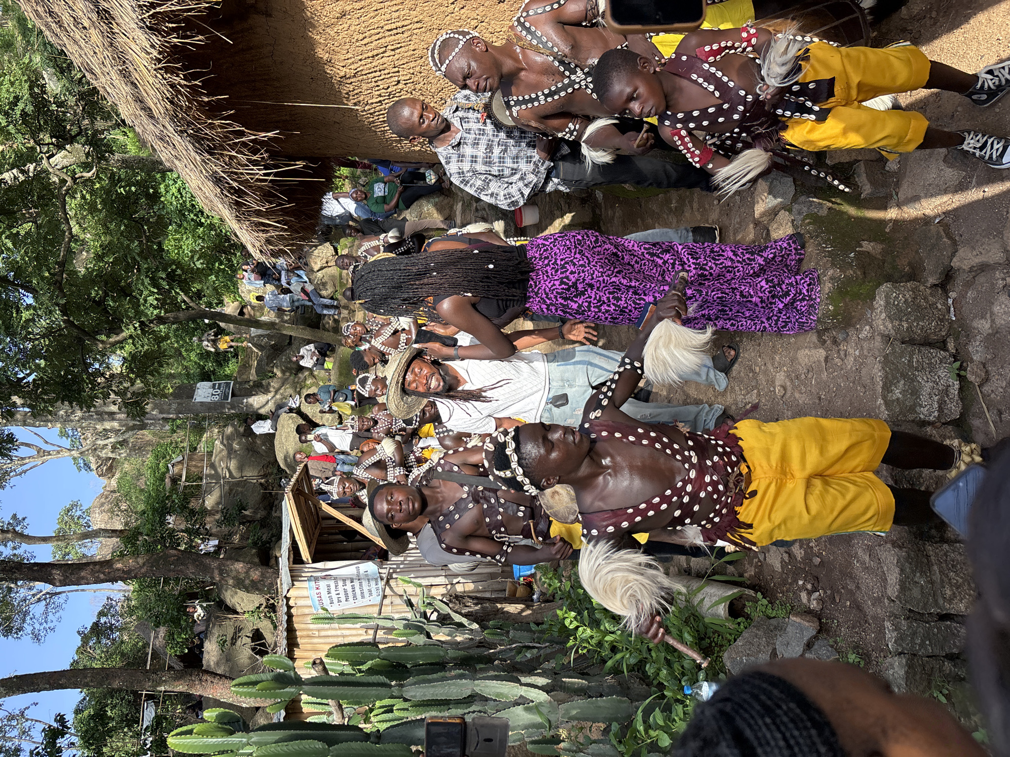 This media file captures a vibrant cultural gathering in Jos, Nigeria. The scene unfolds outdoors, amidst a backdrop of rocky terrain and lush greenery, suggesting a rural or semi-rural setting. A prominent thatched roof structure partially frames the upper portion of the image, hinting at a traditional architectural style.
The central focus is a group of people, many adorned in traditional attire. Several individuals are dressed in striking outfits consisting of patterned vests, shell necklaces, and bright yellow shorts. Some wear white headbands adorned with similar shell embellishments, while others have distinctive white, feather-like ornaments. One person wears a flowing purple skirt with an intricate pattern. The expressions on their faces range from engaged and focused to smiling and joyful, indicating a lively and celebratory atmosphere.
The activity appears to be a cultural performance or ceremony, with the dressed individuals likely participants. The presence of a drum suggests musical accompaniment, and the elaborate costumes imply a significant event. A banner with visible text in English is present, listing food items like "Bush Meat," "Dry & Fresh Pepper Sour," and "Chicken Ma," along with other descriptions, possibly indicating a local market or a food stall at the event. Another sign displays "80" and "80-." The time of day seems to be daytime, with ample natural light illuminating the scene. The overall impression is one of cultural pride and community celebration.