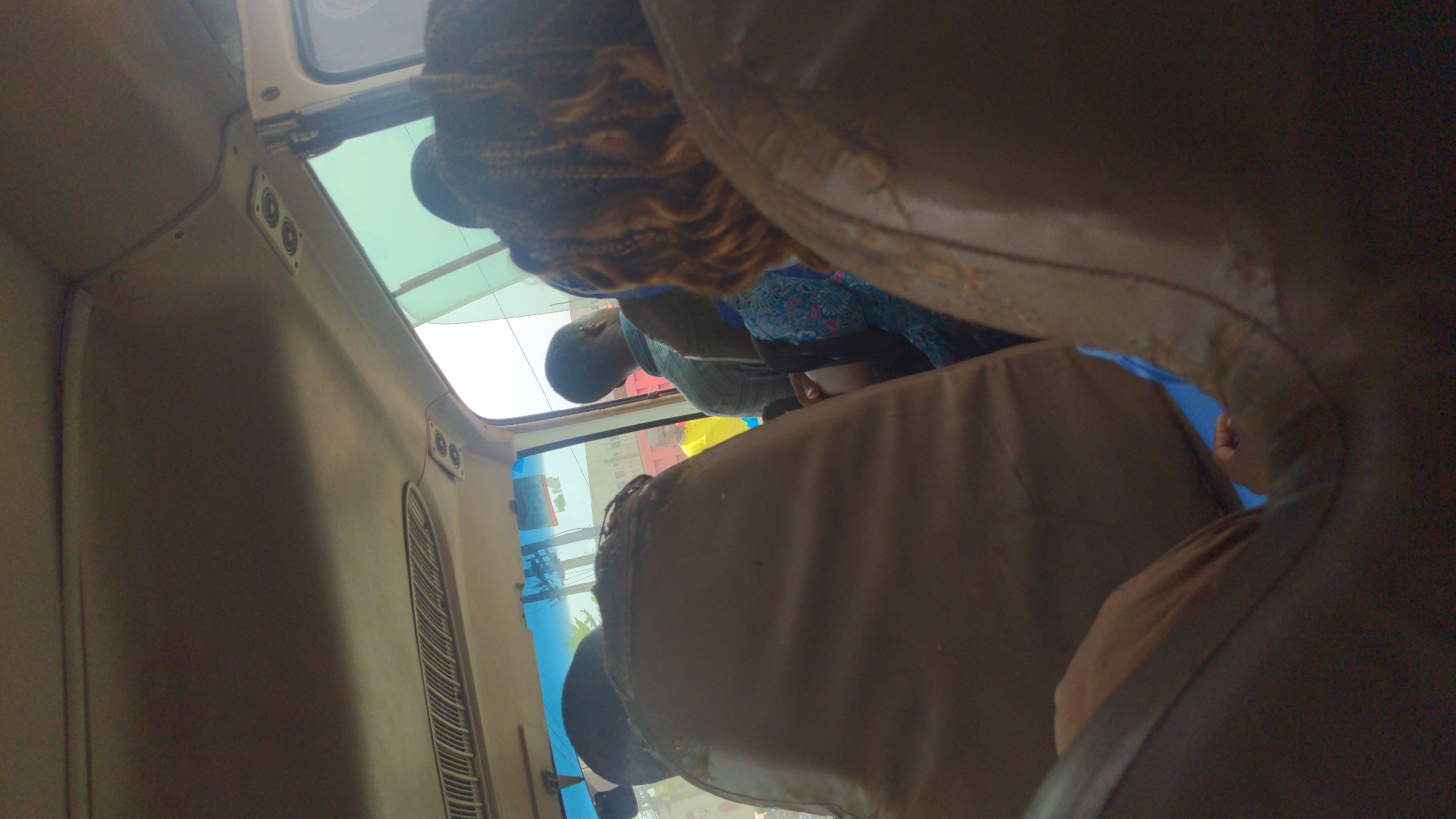 This image, taken inside a passenger vehicle, shows at least three people. A person with braided hair is visible near the vehicle's ceiling. Another person is seated, and part of their head and shoulders are visible. An additional individual is partially obscured by the back of a seat. The vehicle is in Ado, Nigeria, as evidenced by the view through the window, showing an urban environment. The interior of the vehicle appears utilitarian, with worn seats and basic fittings. The vehicle's window is partially visible.