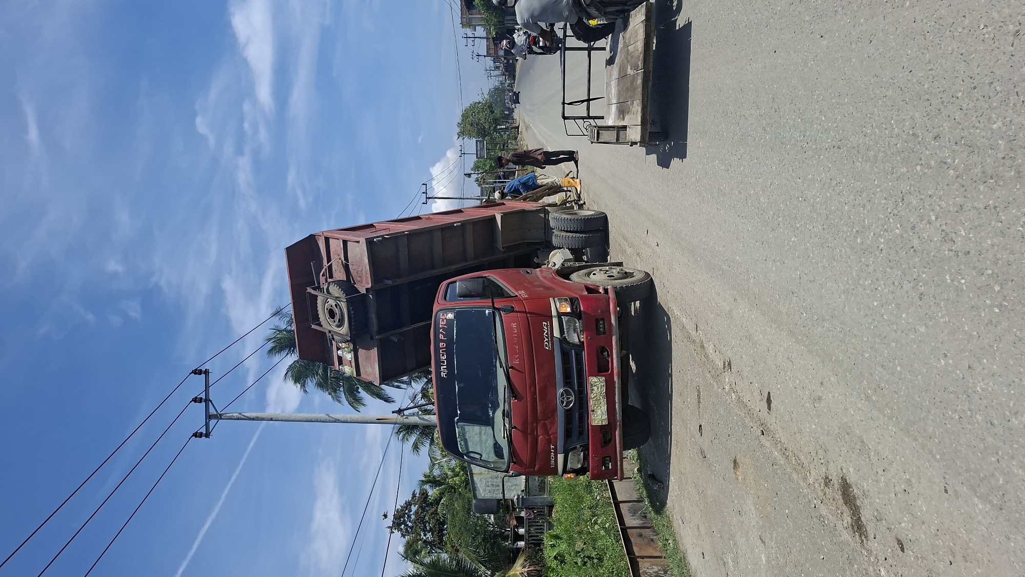 This is a photograph taken outdoors in Beurandang, Indonesia on a bright, sunny day with a partly cloudy sky. The main subject is a red dump truck with its bed tilted upwards, positioned on a paved road. Two men are visible near the rear of the truck. One man, wearing a blue shirt and orange boots, appears to be interacting with the truck's mechanism or the ground beneath it. Another man, in a brown shirt, stands nearby, observing. To the right of the dump truck, a motorcycle with a trailer is parked, and a person is seated on the motorcycle. Further down the road, other vehicles, including cars and motorcycles, are visible in the distance, suggesting ongoing traffic. The environment includes a grassy embankment to the left of the road, lined with vegetation and palm trees. Electrical poles and wires are strung across the scene, typical of urban or semi-urban infrastructure. The road surface is asphalt with some visible patches and minor debris. The overall impression is a street scene with a construction or maintenance activity involving the dump truck.