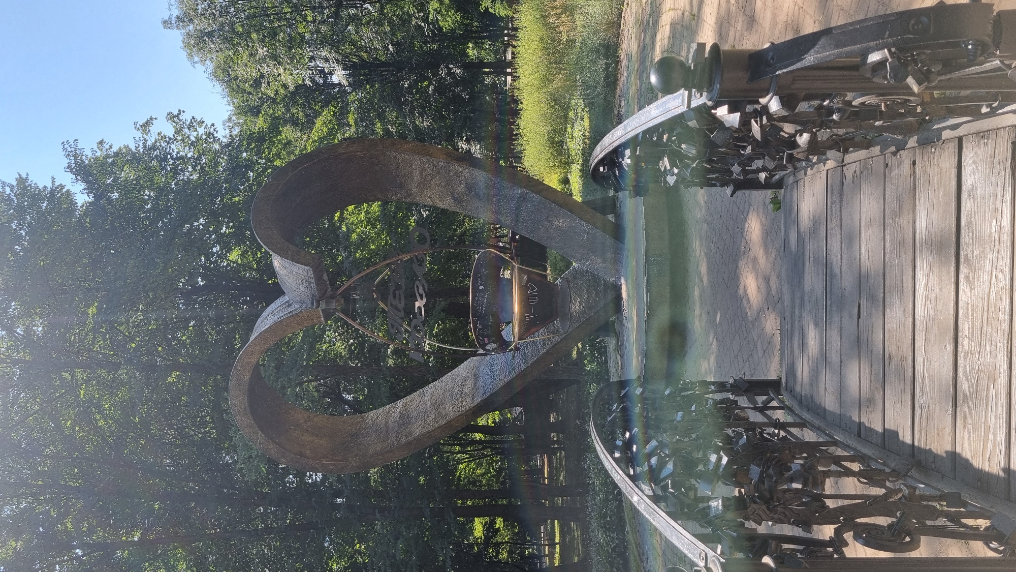 This is a vertical, eye-level, medium shot of a large, bronze heart-shaped sculpture situated outdoors in a park-like setting. The sculpture is positioned centrally in the frame, behind a wooden bridge adorned with numerous padlocks on its railing, a common symbol of commitment. The heart sculpture has an opening in the center, where a darker, bell-shaped object with an inscription is suspended. The background consists of lush green trees and foliage, with a clear blue sky overhead. The lighting suggests it is a sunny day, with some lens flare visible. The overall scene is peaceful and romantic, likely a tourist attraction or a designated spot for couples. This sculpture is located in the city of Chernivtsi, Ukraine.