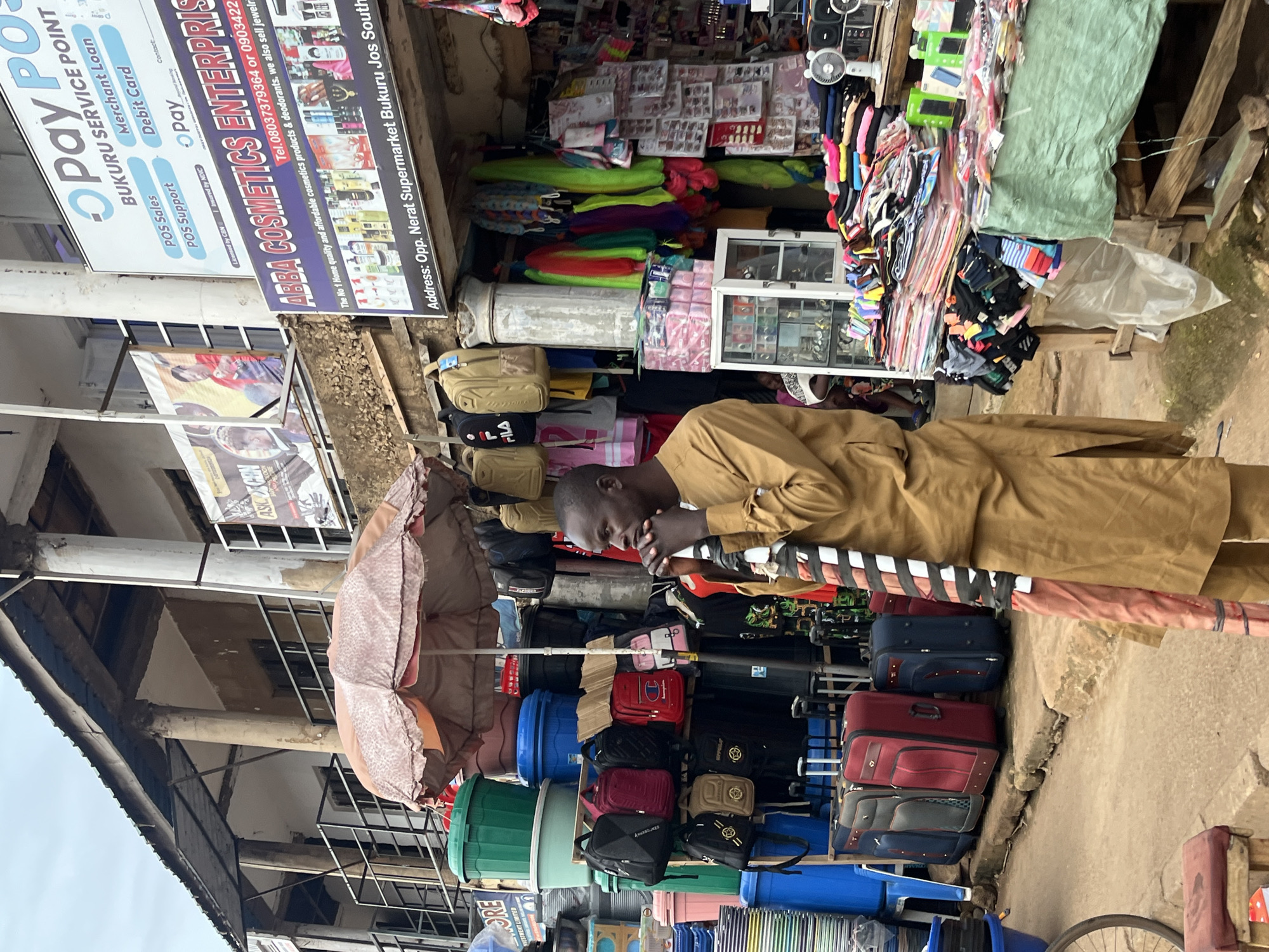 The image depicts a street-level view of a market scene in Bukuru, Nigeria. A man, dressed in a brown tunic and trousers, is positioned in the foreground, facing the right side of the frame with his hands clasped near his mouth. He appears to be holding a striped object, possibly an umbrella or fabric.
Behind the man, the market is bustling with activity. Various stalls are visible, displaying a wide array of goods. To the left, stacks of plastic containers in green and blue hues are prominent, alongside an umbrella providing shade. Further back, several backpacks are hung on display.
The central area of the image features a stall with numerous cosmetic products, as indicated by signage that reads "ABBA COSMETICS ENTERPRISES." The address provided on the sign is "Opp. Nerat Supermarket Bukuru Jos South." Above this stall, a larger banner for "Pay POS SERVICE POINT" is visible, advertising services like "Merchant Loan," "Debit Card," "POS Sales," and "POS Support."
To the right, more market stalls are visible, exhibiting colorful fabrics, folded clothing items, and what appear to be accessories. A small white display cabinet with glass doors showcases smaller items. Several suitcases are neatly arranged on the ground in the lower left portion of the image. The overall environment suggests a busy, open-air market with a mix of goods and services being offered. The architecture in the background shows a building with columns and a corrugated iron roof. The sky is overcast.