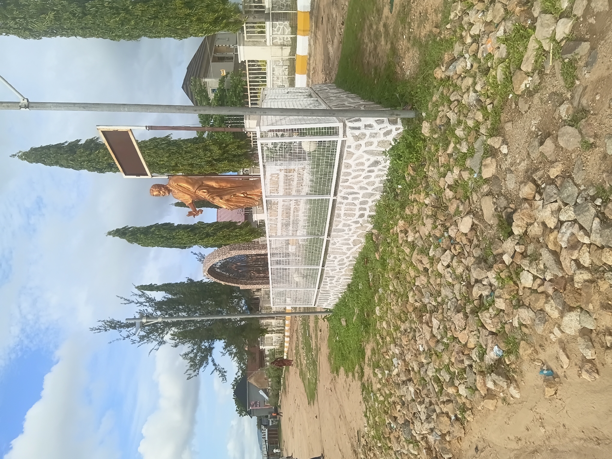 This image depicts a public space or roundabout in Jalingo, Nigeria, under a partly cloudy sky. The central feature is a raised, semi-circular monument base constructed from light-colored, irregularly shaped stones with white mortar, topped by a white metal grid fence.
On this elevated platform stands a golden-colored statue of a robed male figure, holding an object (possibly a scroll or document) in his left hand. To the left of the statue, an ornate, stone-textured archway with dark metalwork is visible. Below the statue, a white panel with partially legible black text includes the word "JALINGO". Above and slightly behind the statue, a blank brown rectangular sign with a white frame is mounted on a pole. Several tall, slender evergreen trees are positioned behind and around the monument.
The ground in the foreground and to the left consists of a mix of sandy dirt and numerous scattered rocks, with patches of green weeds and some discarded plastic litter. A dirt path or road runs from the foreground into the background on the left. On the right side, a short section of yellow and white painted curb marks the edge of the path near the monument.
In the background, multiple buildings are visible, including a modern-style house with a brown roof and white walls on the right, and other structures further in the distance. Several individuals are present on the dirt path on the left; one person in a patterned dress is walking away from the viewer, while others are further in the distance. The scene is illuminated by natural daylight.