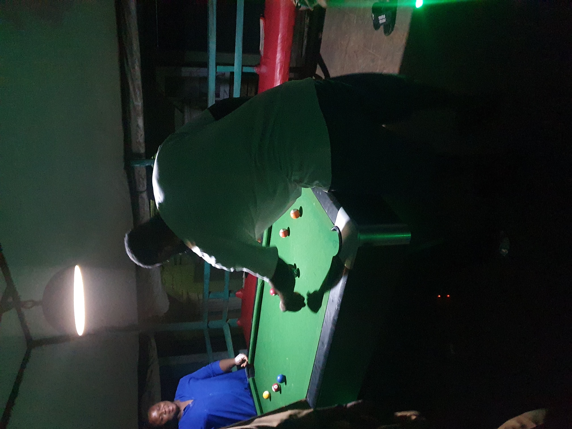 In Lokoja, Nigeria, a dimly lit indoor space features a green billiards table. Two individuals are visible near the table. One person, wearing a blue shirt, stands on the left, appearing to be observing. The second person, on the right, is actively engaged in playing, with their back partially to the camera, wearing a light-colored t-shirt and dark pants. Several billiard balls are scattered across the table. The environment includes overhead lighting, a red bench to the right, and metal railings. The overall atmosphere is informal, suggesting a recreational setting.