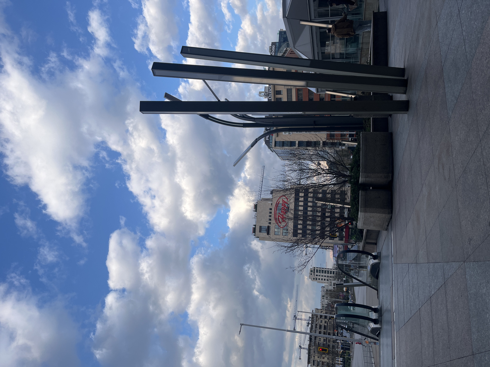 The image depicts a cityscape in Sofia, Bulgaria, identifiable by the "Happy" restaurant sign on a building. The foreground shows modern architectural elements, including square pillars and curved metal structures, possibly part of a building's facade. There are also square planters. The cityscape includes multiple buildings, some under construction with cranes visible. The sky is partly cloudy with patches of blue. No people are clearly visible, though indistinct figures are present behind the architectural elements. The environment is urban, with signs of construction and commercial activity.