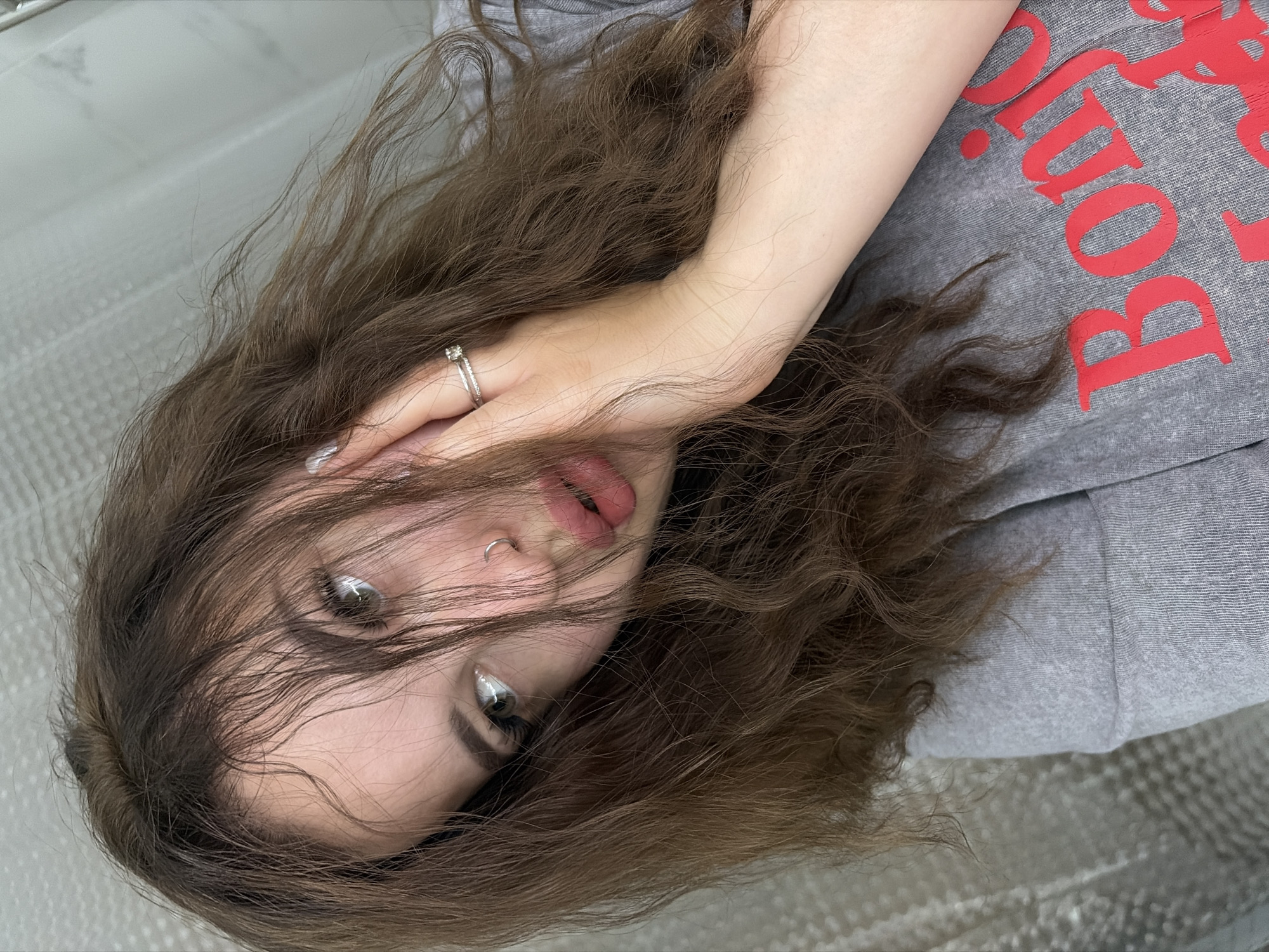A young woman is depicted in a close-up, slightly tilted portrait. She has long, wavy brown hair that partially obscures her face. Her eyes are visible, and she has a silver ring in her nose. She's wearing a gray sweatshirt with a large red inscription, with a ring worn on her finger. The background appears to be a textured, possibly shiny surface, which could be flooring or a similar material. The setting or location of Krasnodar, Russia, cannot be determined from the visual information provided.