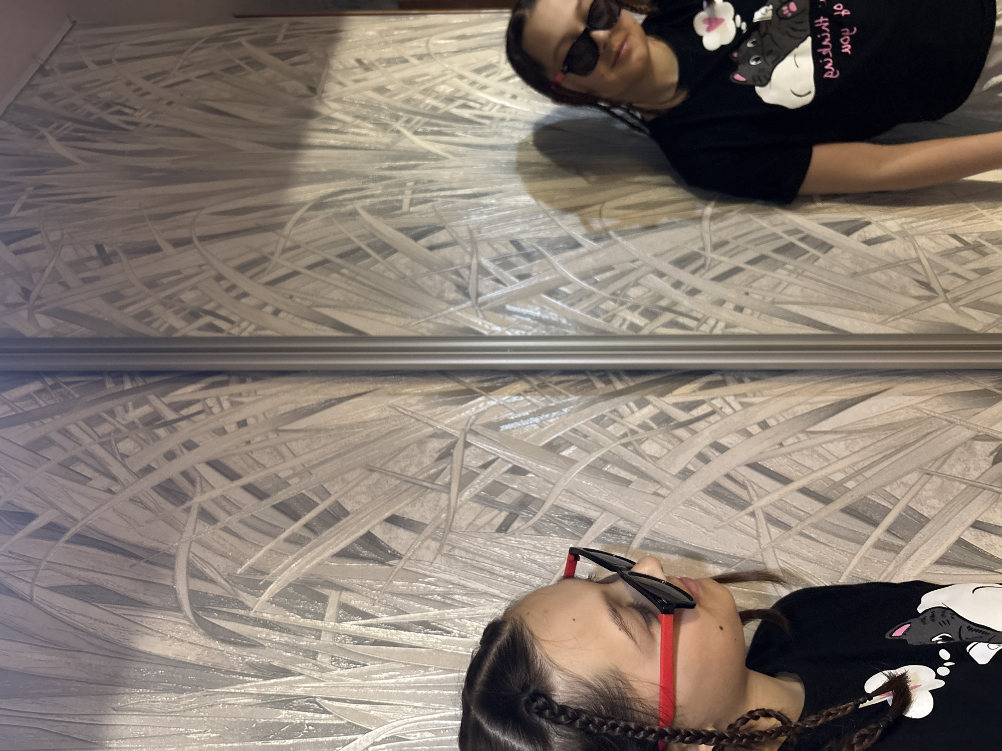 This image depicts a person, likely a young individual, situated in an indoor environment in Chaikovsky, Russia. The scene is horizontally bisected by a reflective surface, possibly a mirror or a wardrobe panel.
In the lower portion of the image, the person is shown directly, lying down. They are wearing black sunglasses with distinct red arms and a black t-shirt adorned with a cartoon design of a black and white cat within a thought bubble. Pink text on the t-shirt reads "thinking of you." The person has dark hair styled in braids.
The upper section of the image presents a reflection of the same individual, appearing mirrored and inverted due to the reflective surface. This reflection also shows the person in a relaxed, lying position.
The background on both sides of the reflective divide features a wall covered with light-toned wallpaper. The wallpaper pattern consists of numerous abstract, intertwined, and elongated shapes, reminiscent of blades of grass or reeds, creating a dynamic visual texture. The lighting suggests an indoor setting. The person's eyes are concealed by sunglasses, contributing to a relaxed demeanor.