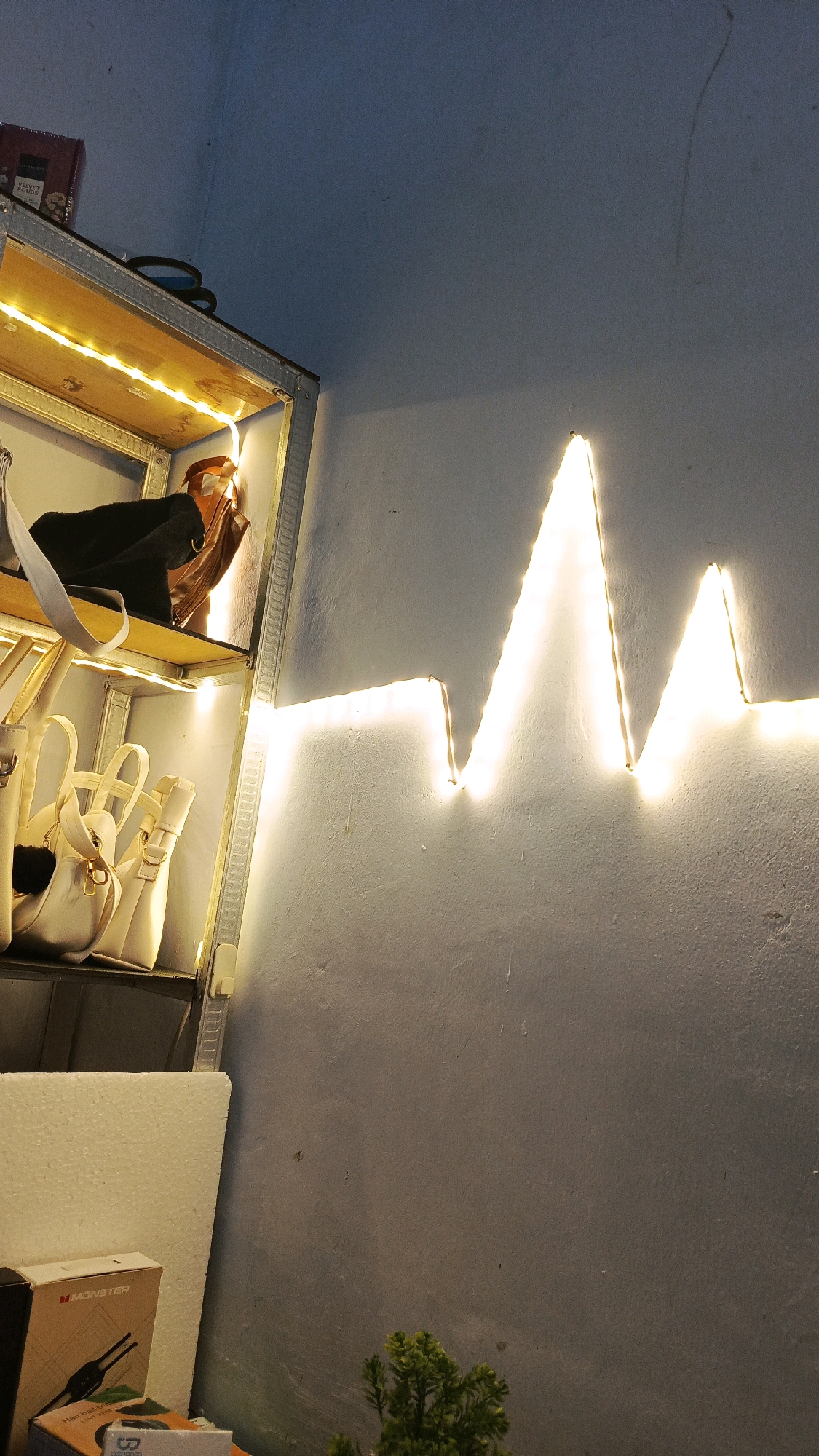 The image depicts an indoor scene with a shelf unit on the left, displaying several handbags and what appears to be a boxed item on the top shelf. The shelf unit is illuminated by warm LED strip lights. On the right side of the image, a wall is decorated with a string of LED lights that form a zigzag pattern resembling a heartbeat or sound wave. The wall has a textured, rough surface, and the lighting creates shadows and highlights. In the lower left corner, there are boxes and what appears to be a piece of styrofoam. A small, green artificial plant is visible in the lower right corner. The overall lighting suggests a low-light environment. The scene is likely an interior space, possibly a shop or a home. The city of Babadan, Indonesia is not explicitly depicted or suggested by any elements within the image.
