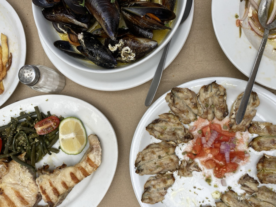 The image displays a forensic overhead view of a dining table in Skala, Greece, laden with several white plates of partially consumed food. At the top-left, an oval plate contains a grilled fish fillet exhibiting distinct sear marks, a serving of green beans with visible seasoning, a halved cherry tomato, and a sliced lime wedge. Directly below this, a second oval plate holds multiple grilled sardines, some reduced to remnants, surrounding a central pile of diced red tomatoes and red onions. A silver fork rests on this plate. To the right, a white bowl is filled with mussels in a pale yellow broth. A silver spoon is positioned next to this bowl. A portion of a white plate with french fries is visible at the top-right, and another white plate containing a salad with red onions is partially seen at the bottom-right, also with a silver spoon. A fluted clear glass salt shaker stands between the top-left fish plate and the mussel bowl. The entire arrangement rests on a plain, light tan table surface. No individuals are present in the frame.